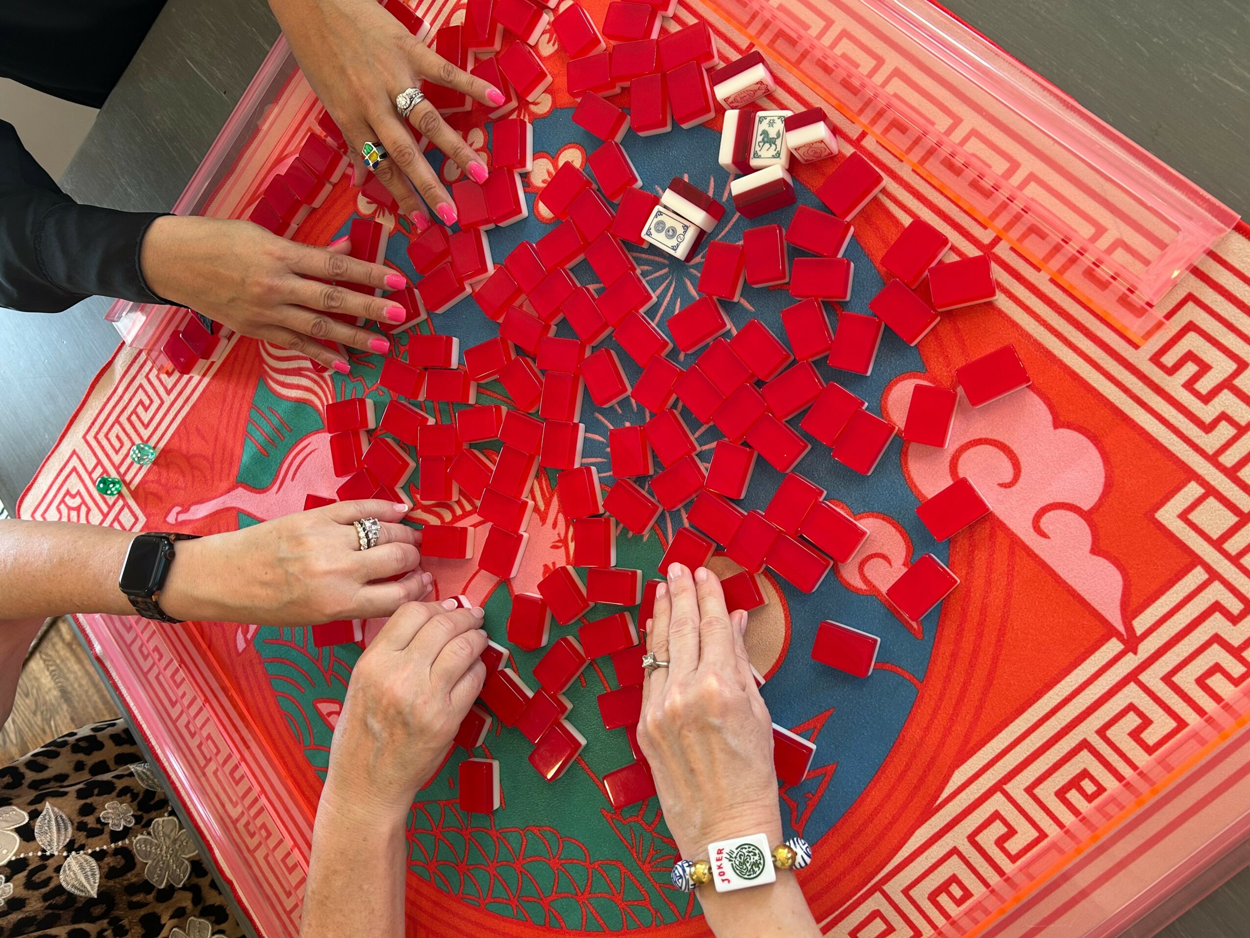 People playing a game with red and blue tiles on a red patterned table, hands reaching for tiles.