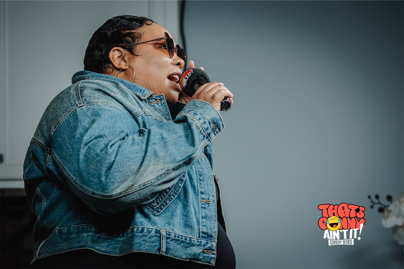 Person singing into a microphone, wearing sunglasses and a denim jacket, against a plain background.