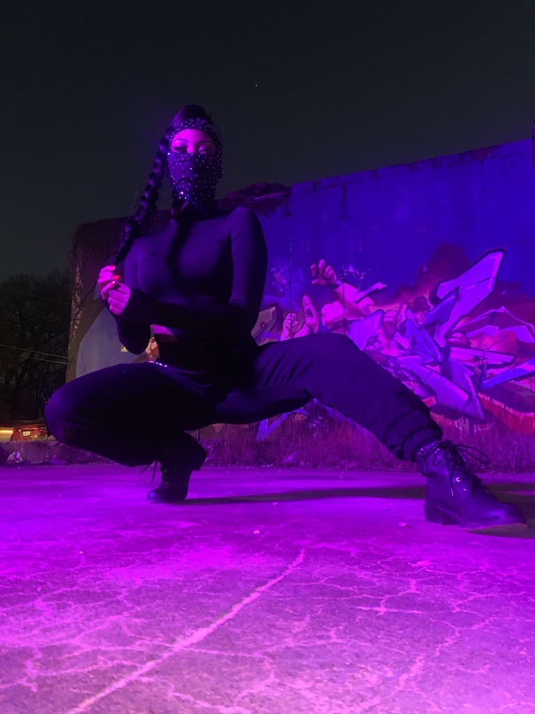 Person crouching outdoors at night, illuminated by purple light, with a mural in the background.