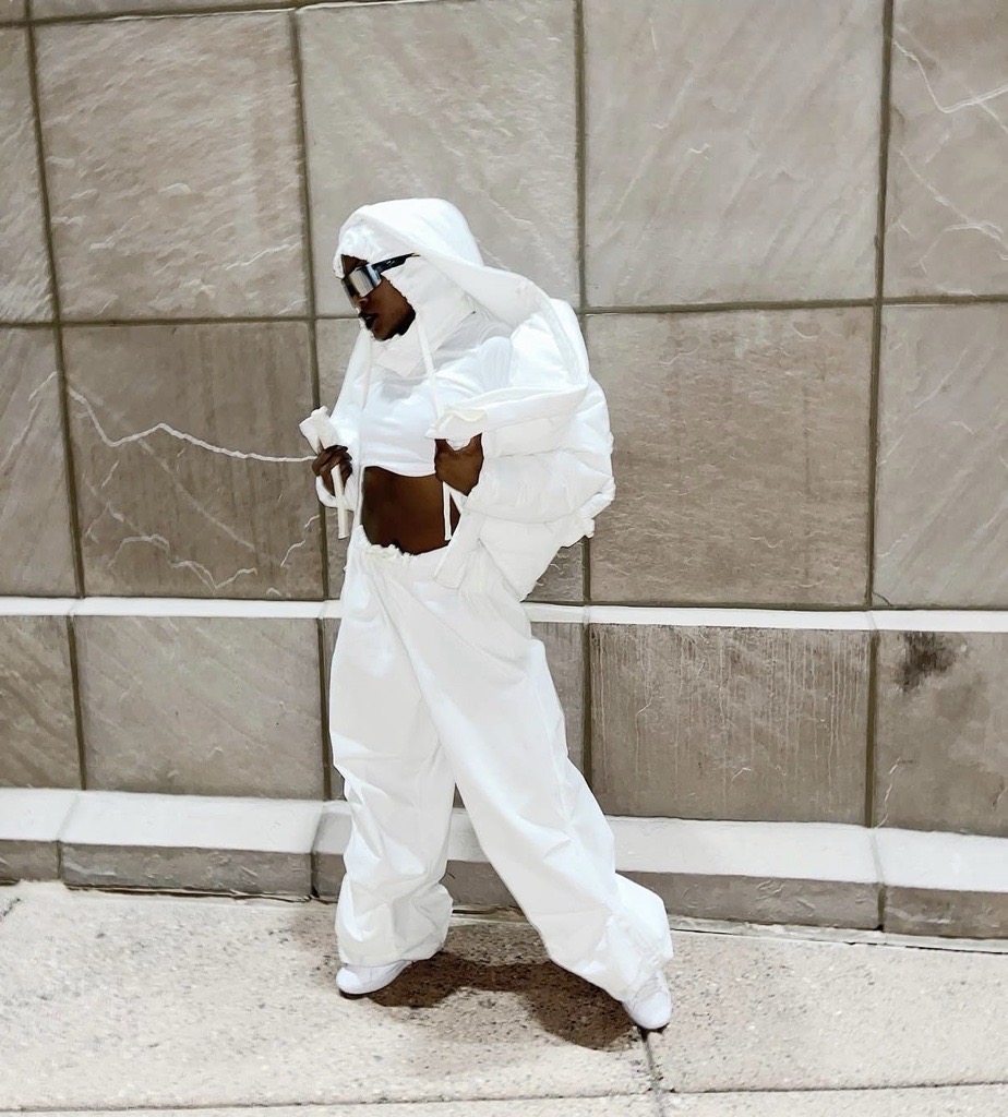 Person in white protective suit and sunglasses standing against a tiled wall, looking at a phone.