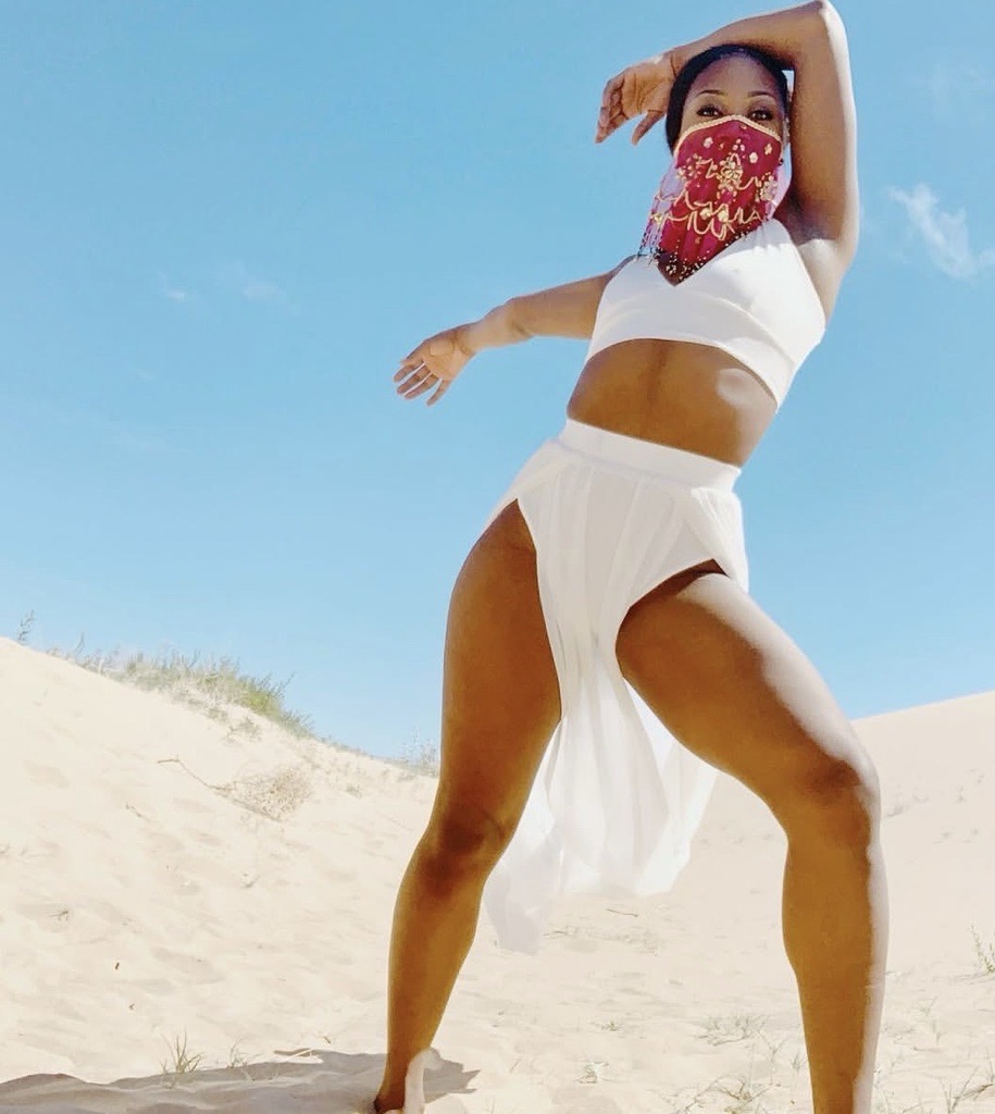 Woman in white outfit with a red face mask standing on sandy beach under blue sky, arms raised, one leg forward.