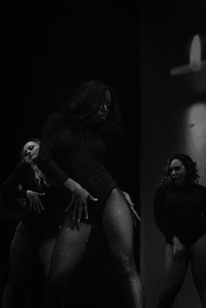 Three women standing in a dark room with a lit candle on the wall, one woman in the center with glittery legs.