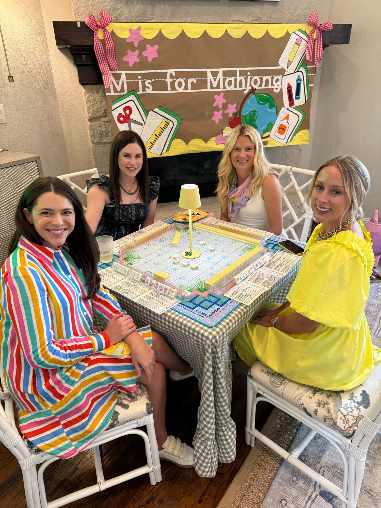 Four women sit around a table playing a board game, with a birthday banner in the background, in a decorated room.