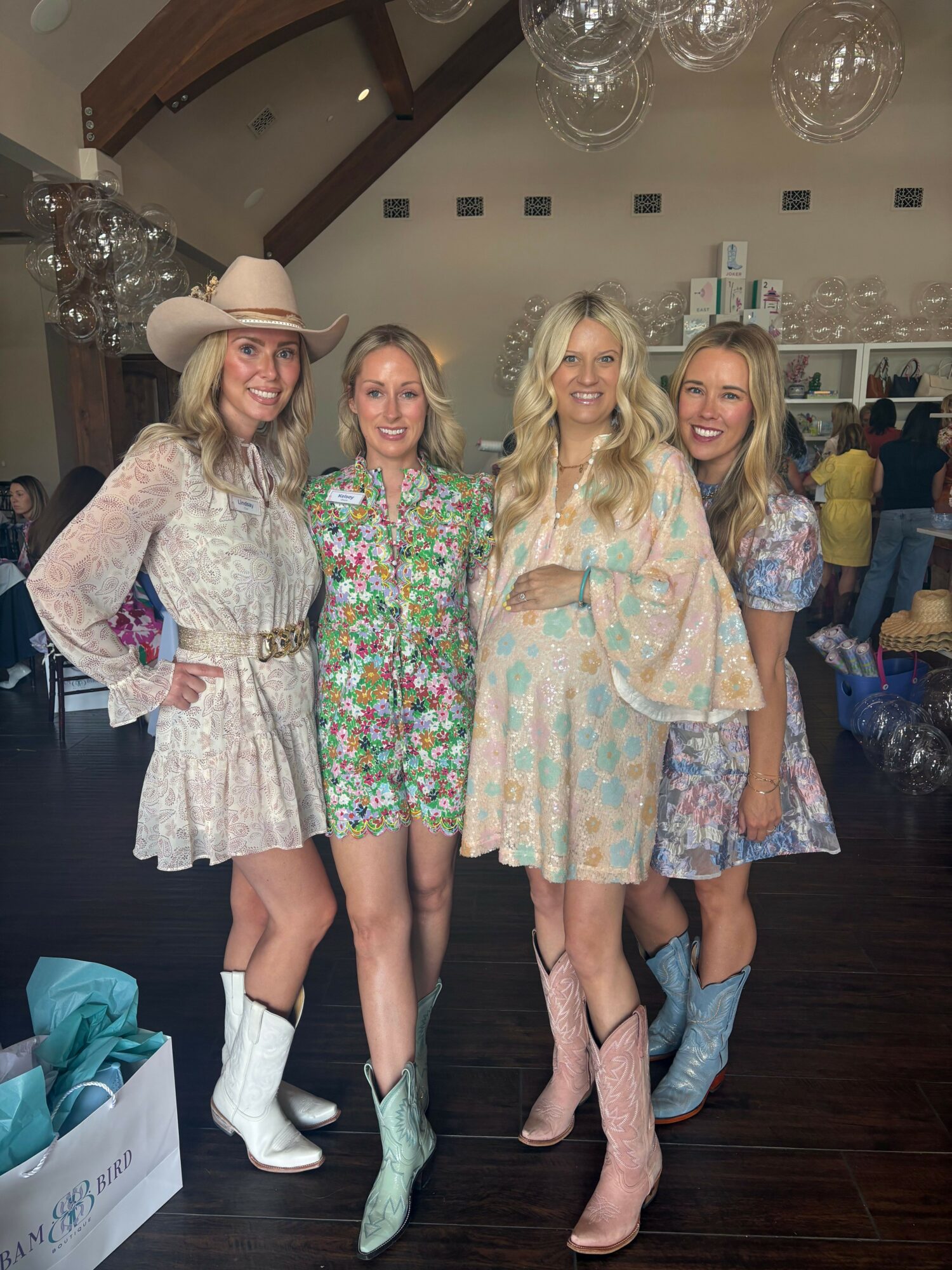 Four women in colorful dresses and boots pose indoors with dark wooden floor and glass chandeliers.