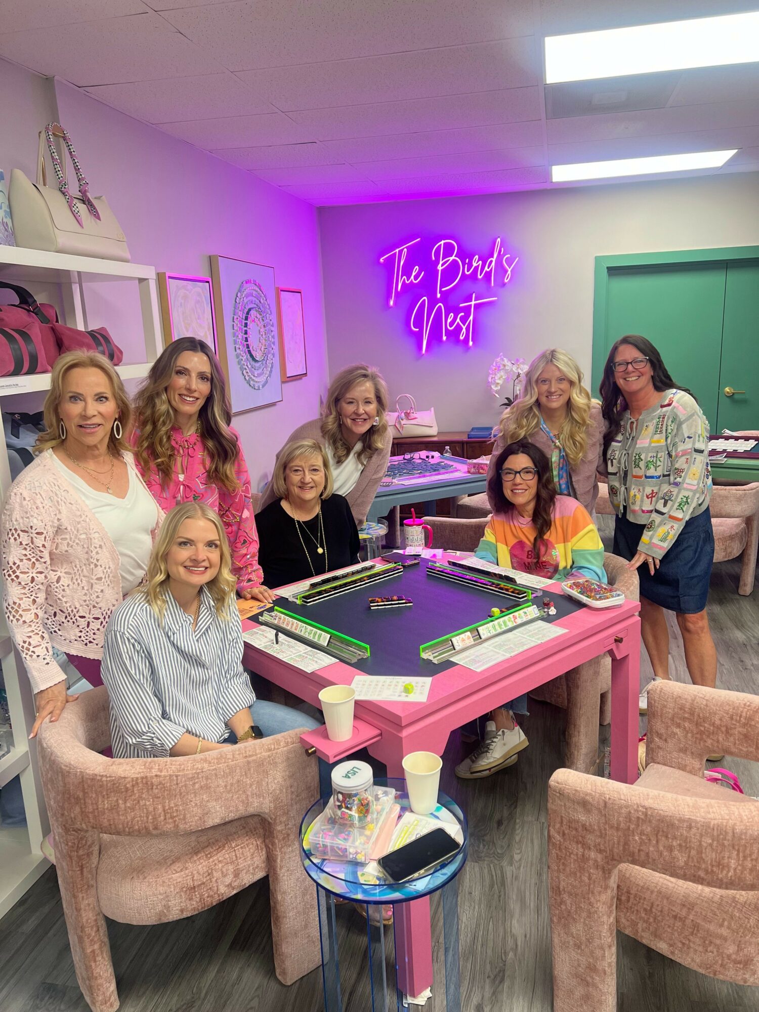 Group of women gathered around a pink pool table in a room with neon sign and artwork on the wall.