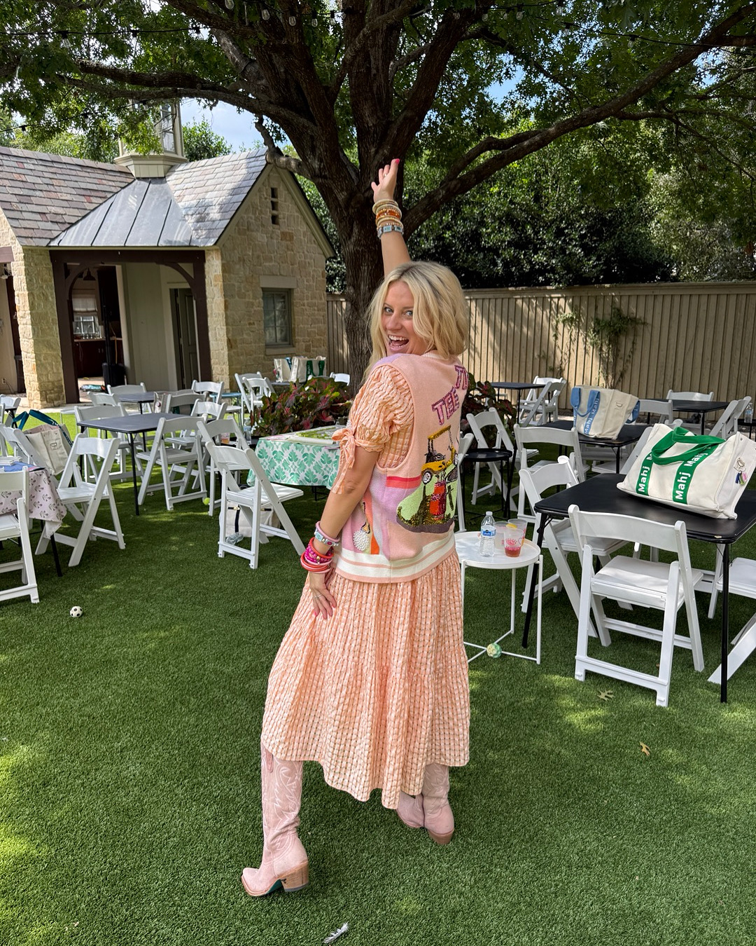 Woman with blonde hair in a peach dress standing outdoors under a tree, smiling, with chairs and tables in the background.