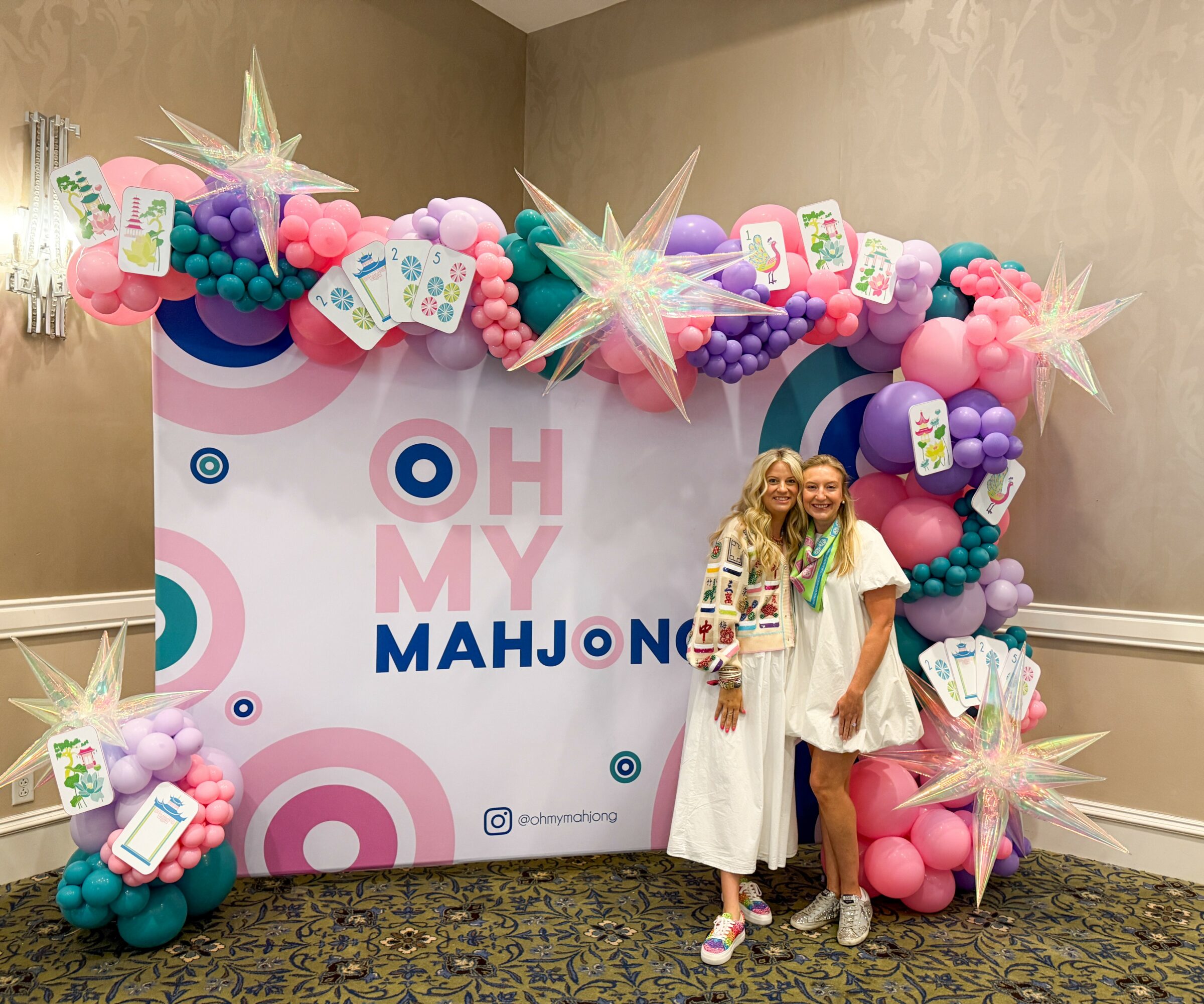 Two women stand in front of a decorated backdrop with pink, purple, and blue balloons, stars, and cards, celebrating a baby shower.