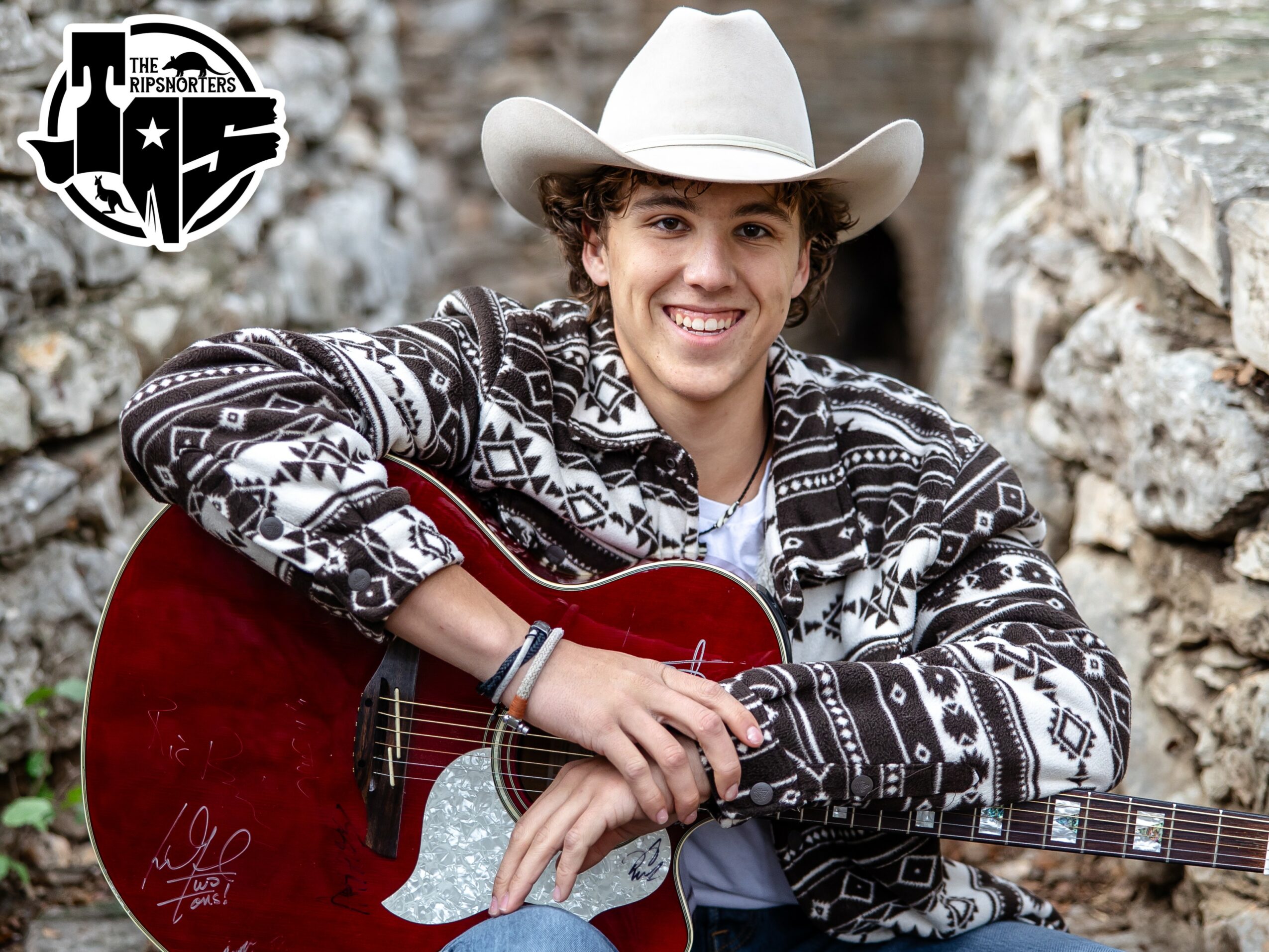 Young person wearing a cowboy hat and patterned jacket, smiling while holding a red acoustic guitar outdoors.