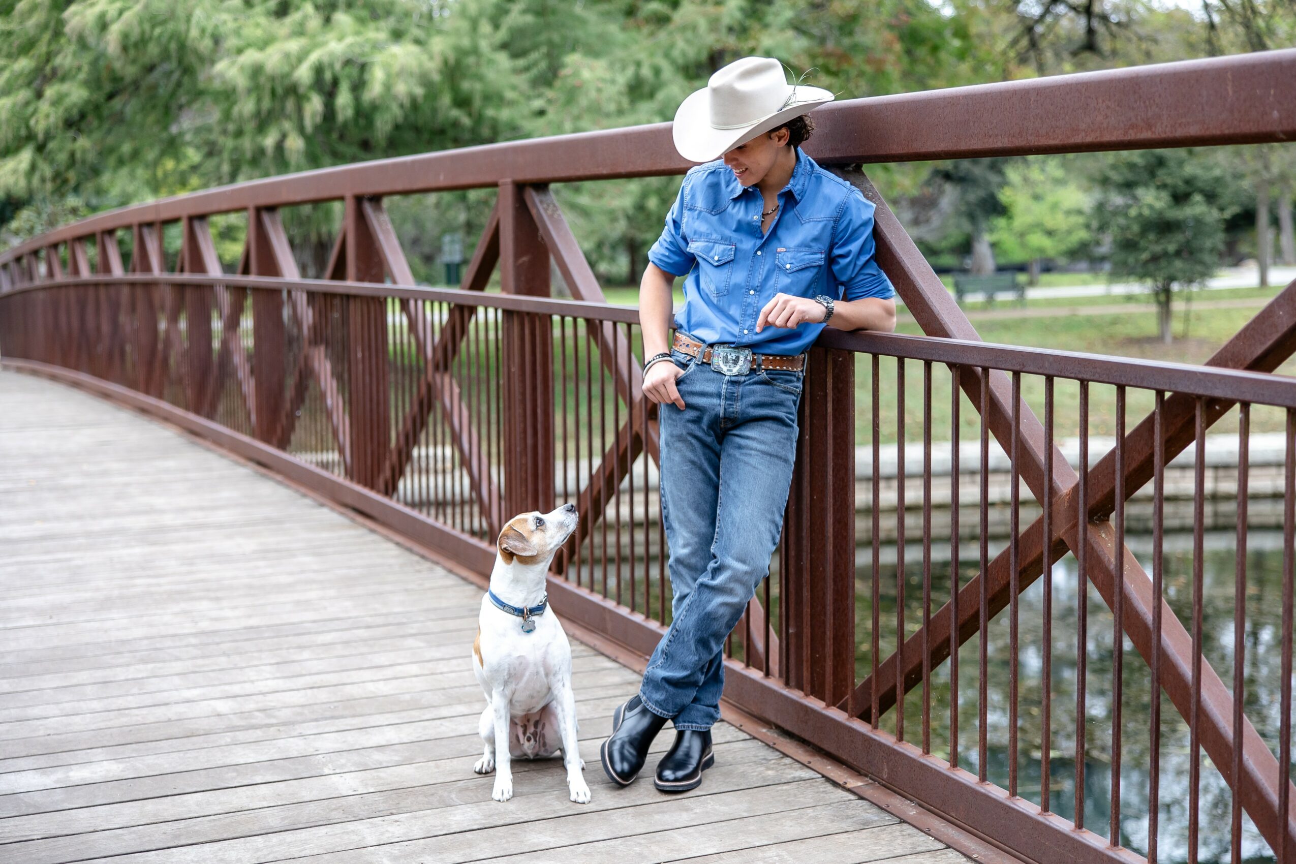 Person in denim outfit and wide-brimmed hat leaning on bridge railing with dog on leash, outdoors in park, trees in background.