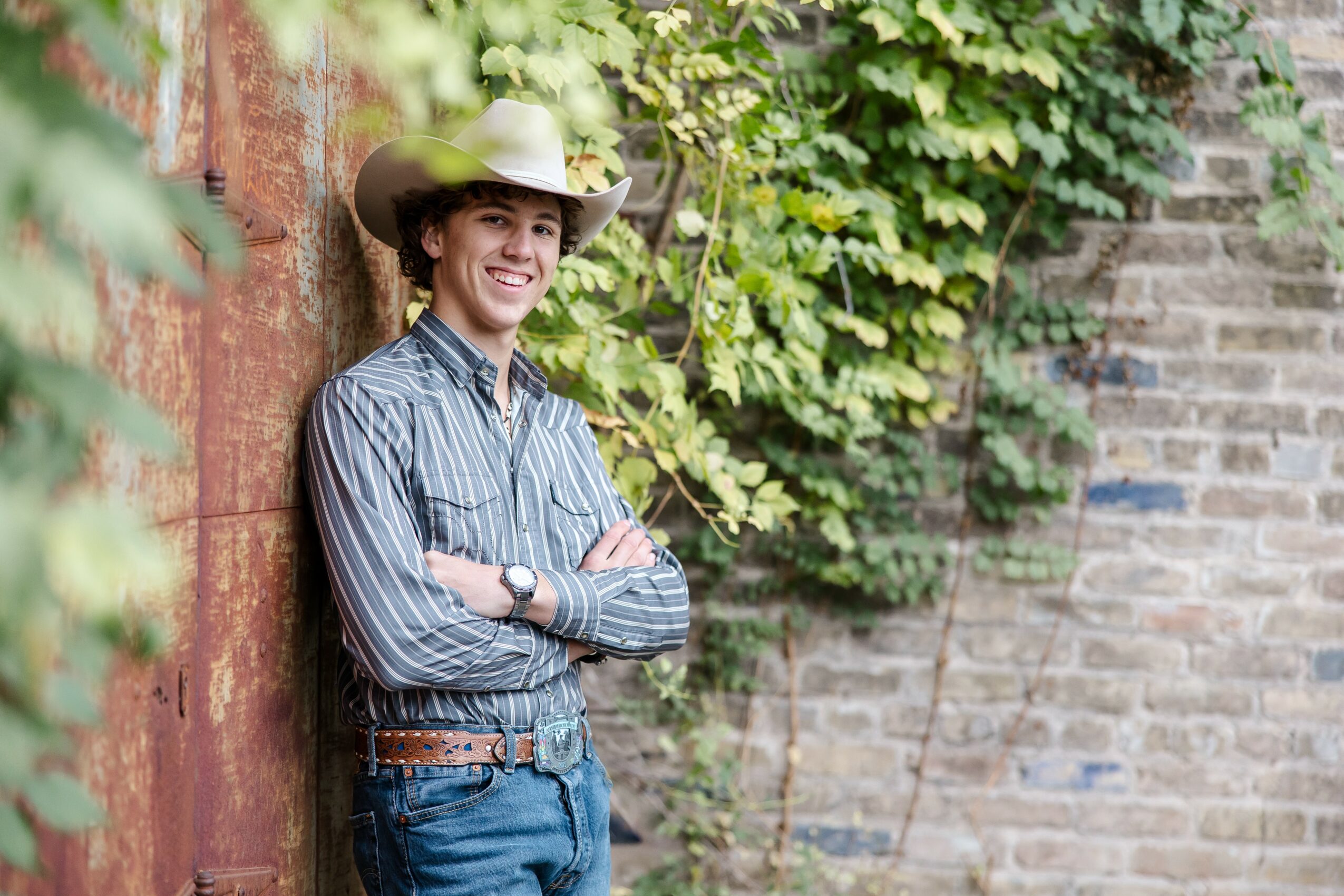 Young person wearing a hat and striped shirt, smiling, standing outdoors near a brick wall and green foliage.