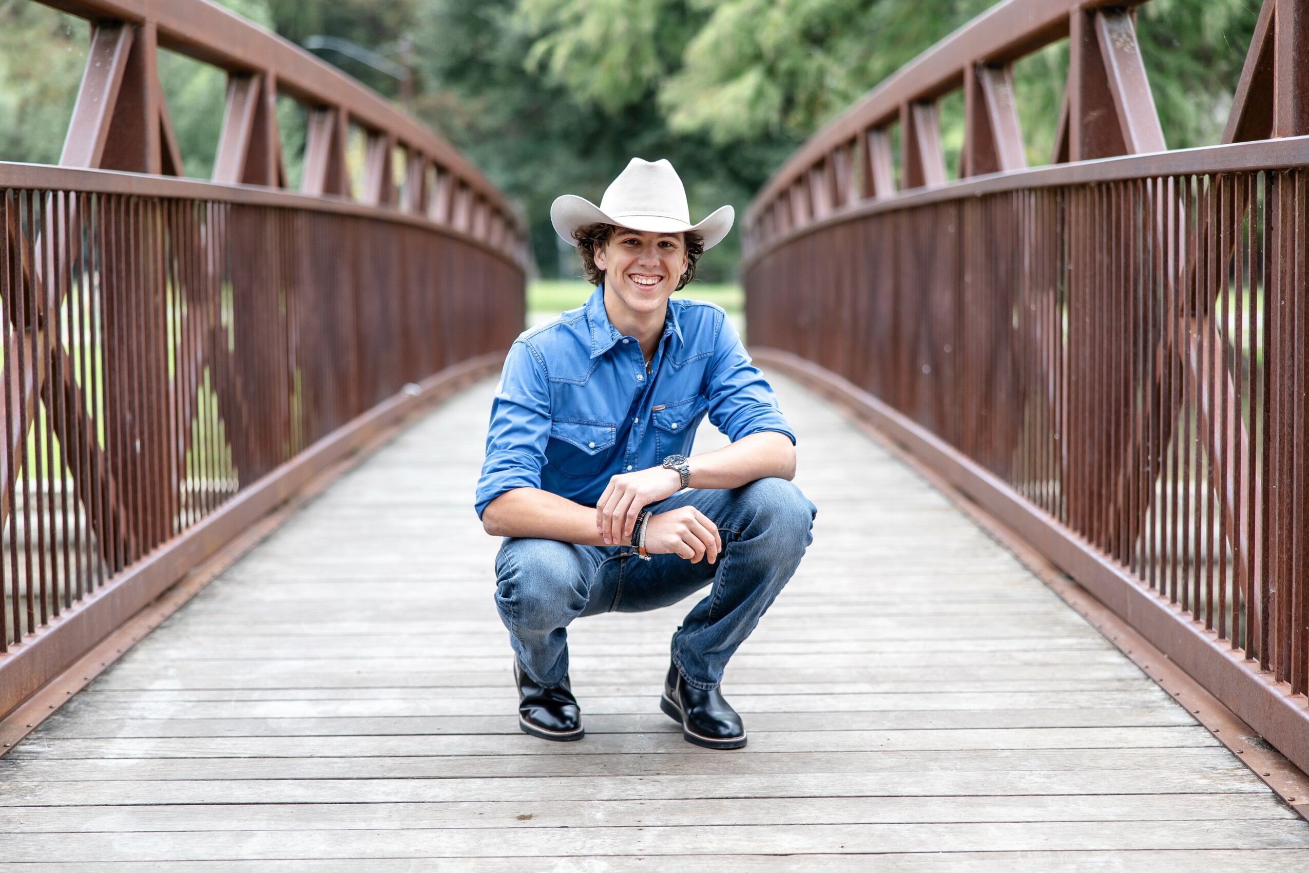 Person wearing a cowboy hat and blue shirt squatting on a wooden bridge with metal railings, outdoors with trees in background