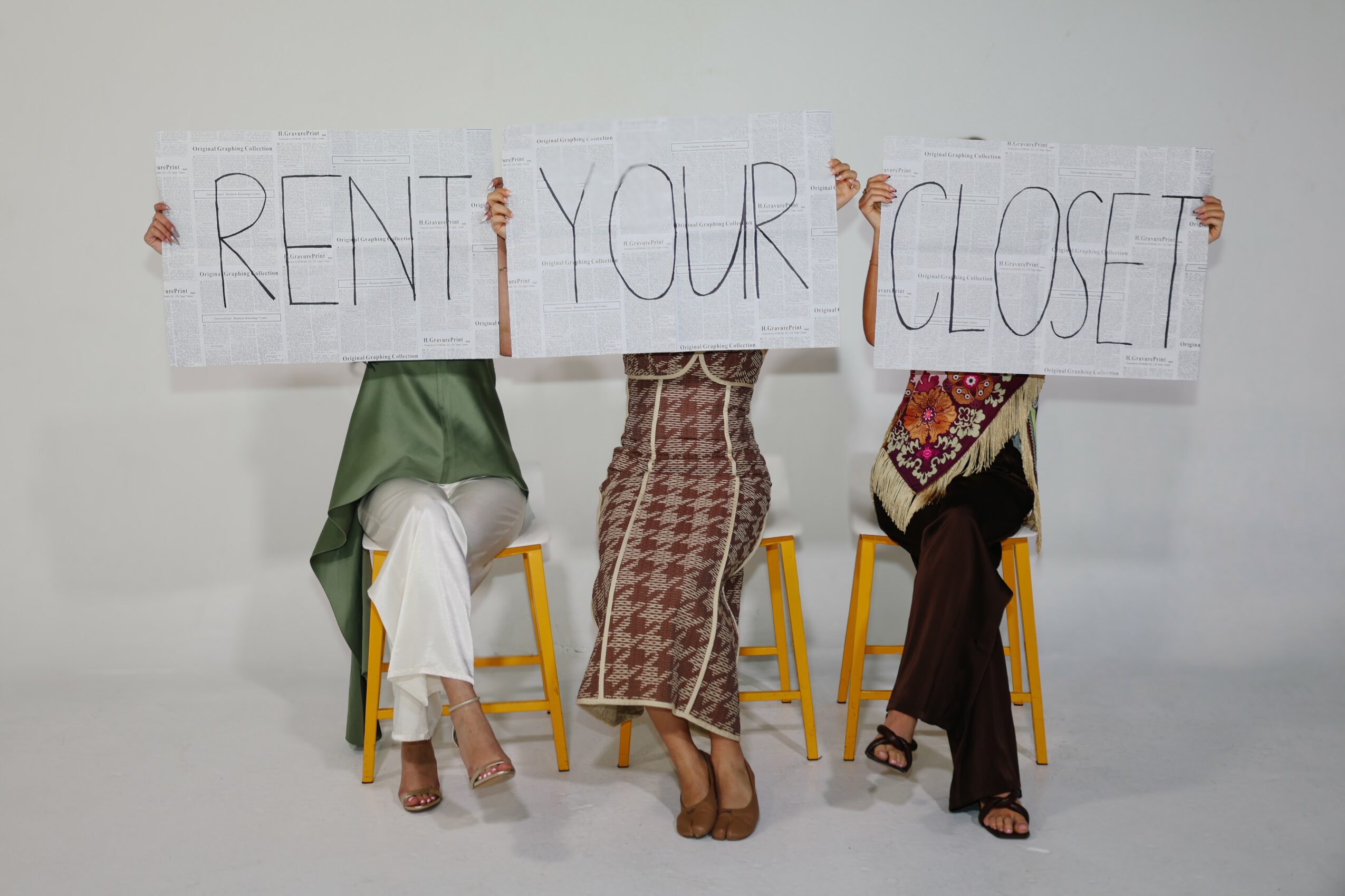 Three people sitting on yellow chairs holding a sign that says 'RENT YOUR CLOSET'.