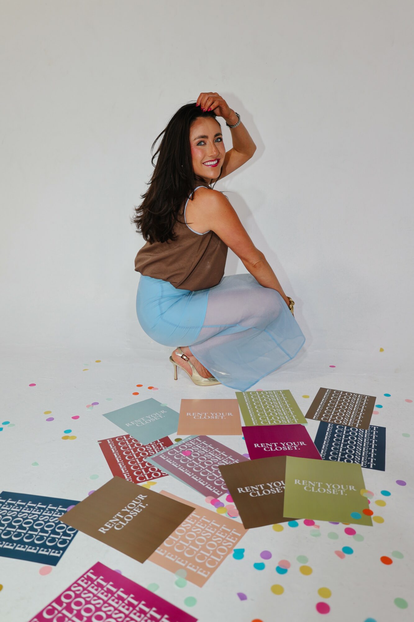Woman crouching with books and colorful confetti on white background, smiling, wearing a brown top and light blue jeans.