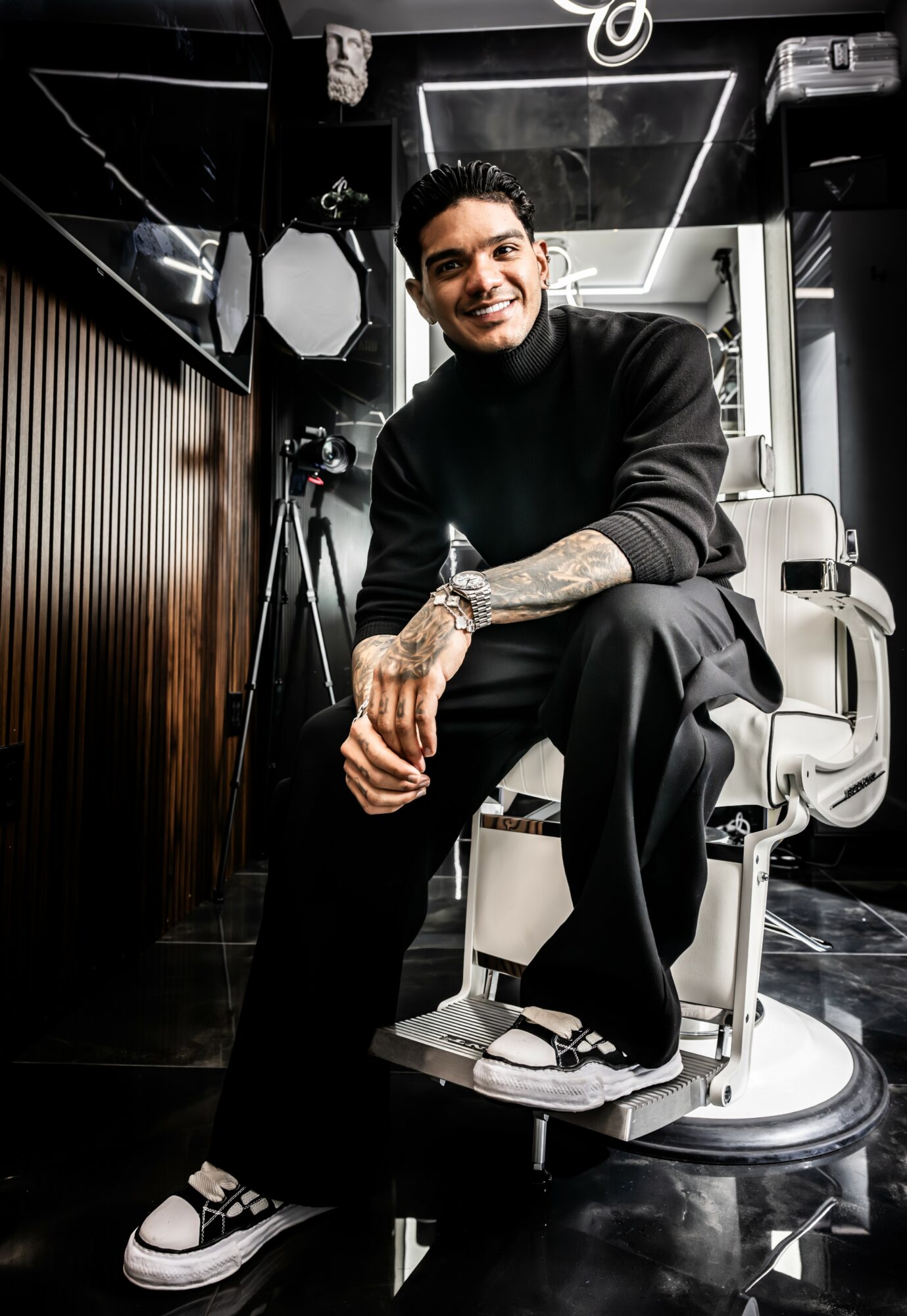 Young man with tattoos smiling, sitting on a barber chair in a modern salon, wearing black clothing and sneakers.