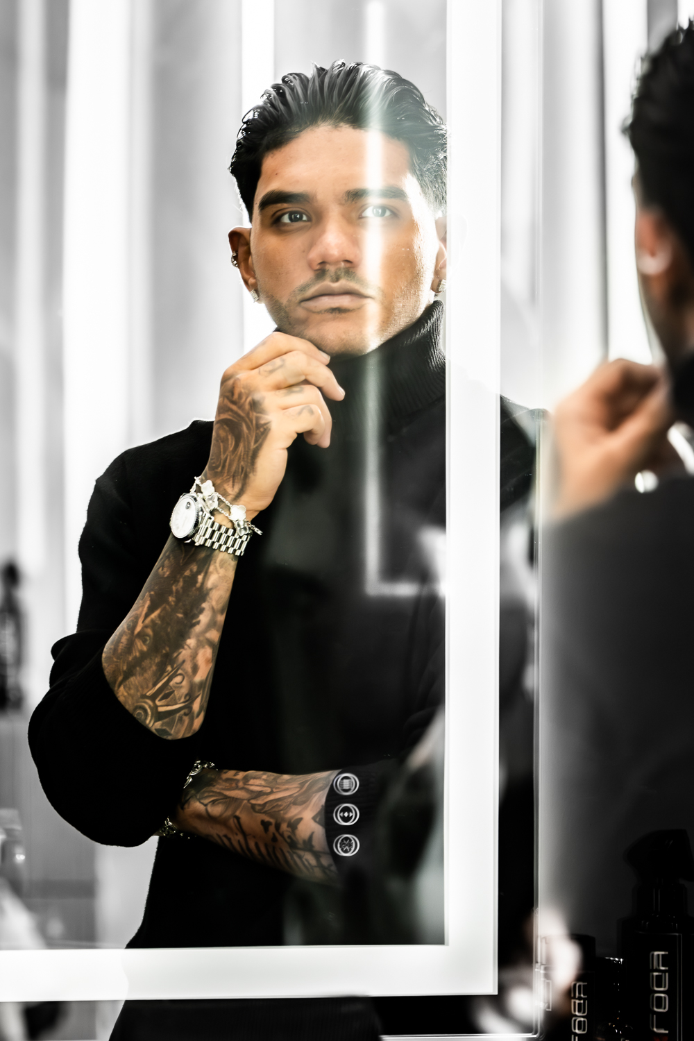 Man looking at his reflection in a mirror, with tattoos on his arm, wearing a black shirt, in a bright room.
