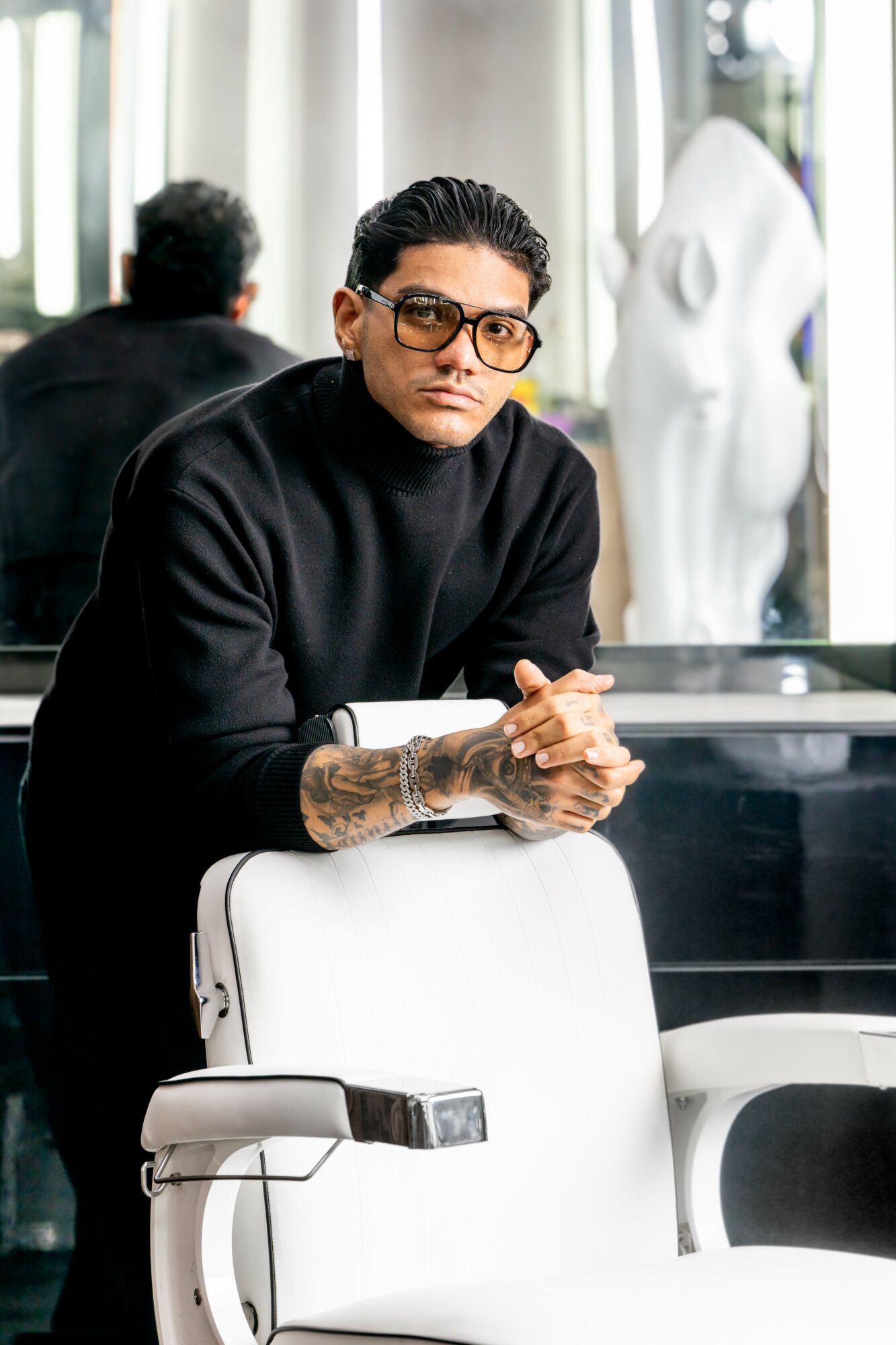 Man with glasses and tattoos sitting at a table in a modern indoor space, with a large sculpture and window behind him.