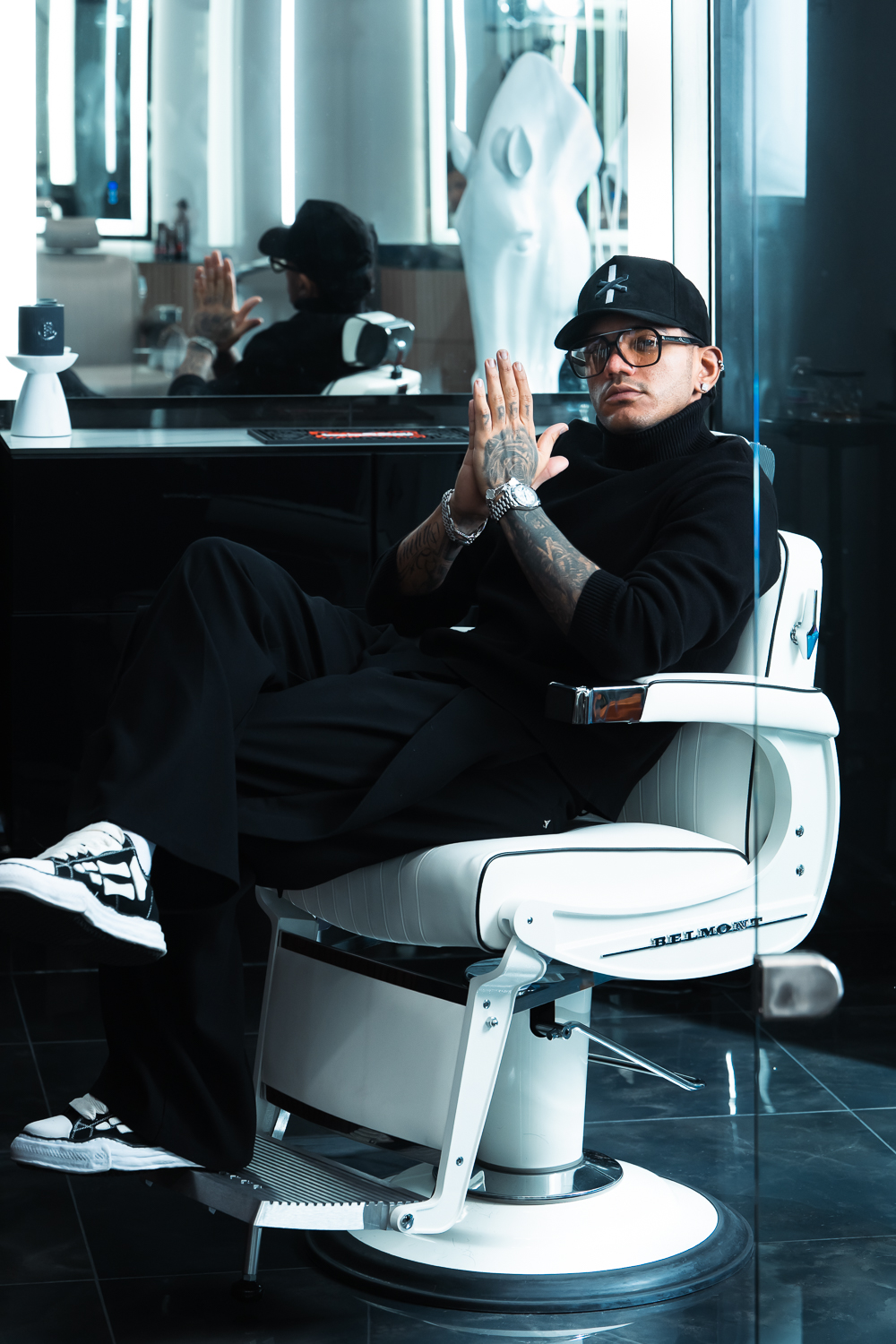 Person sitting in a chair with hands clasped, wearing glasses and a cap, in a modern room with reflective surfaces.