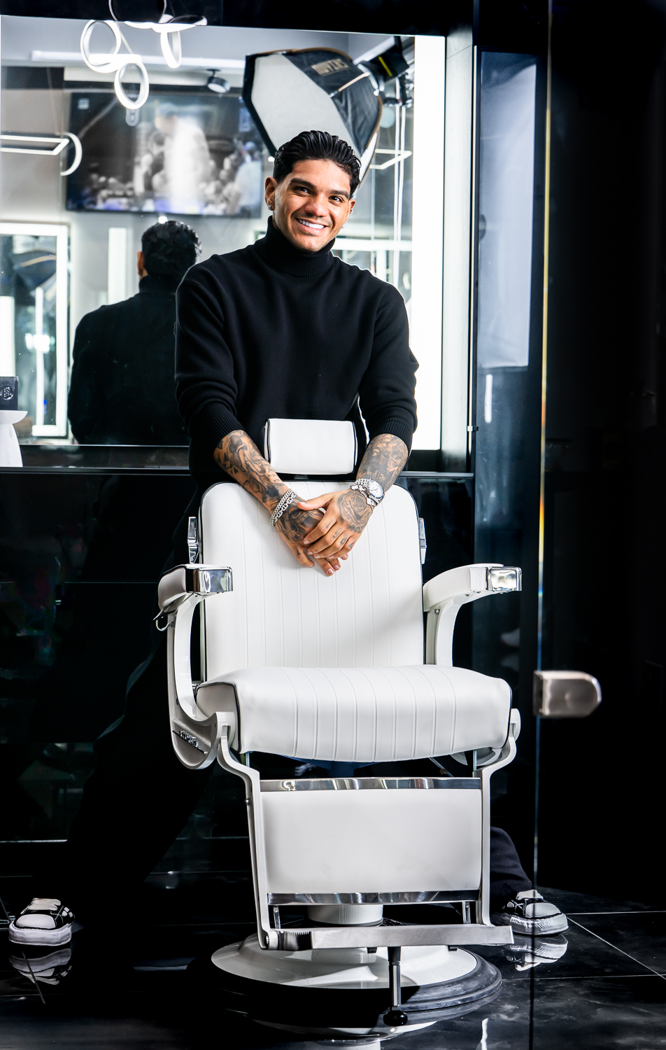 Young man with tattoos smiling, standing behind a white barber chair in a salon, holding a box, with mirrors and equipment in background.
