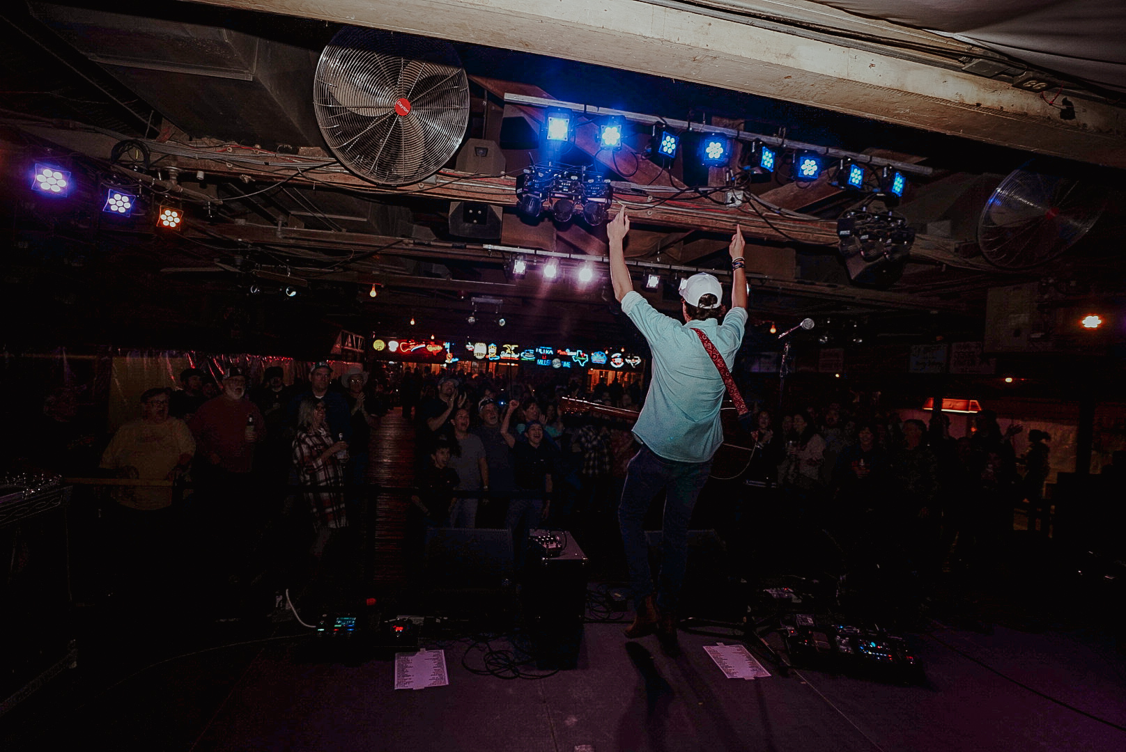 Person on stage raising arms in front of a crowd with stage lights and equipment overhead.