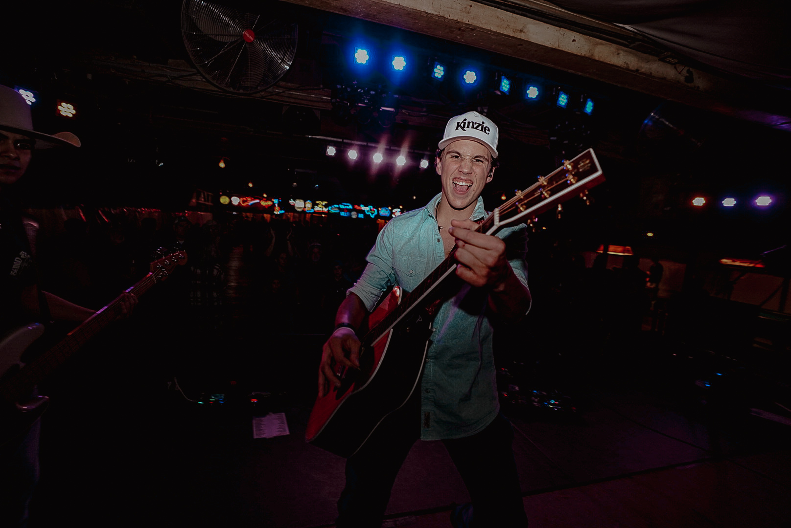 Person playing guitar on stage with colorful lights, smiling, wearing a cap and casual clothing.