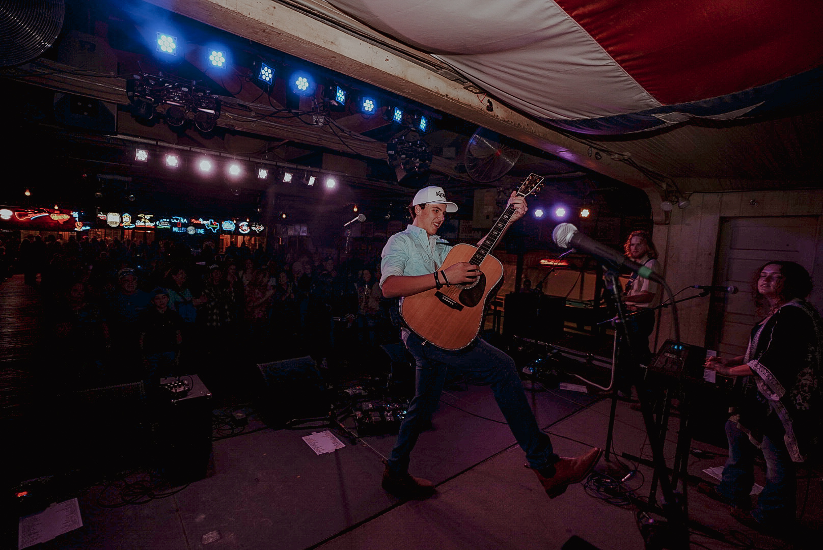 Musician playing guitar on stage with band, colorful lights, audience in background, indoor concert setting.