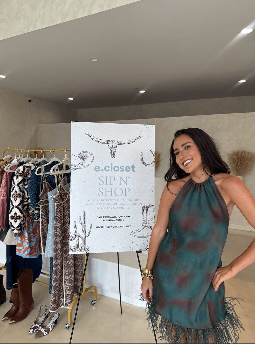 Smiling woman in a sleeveless dress standing next to a clothing rack and a sign in an indoor space.