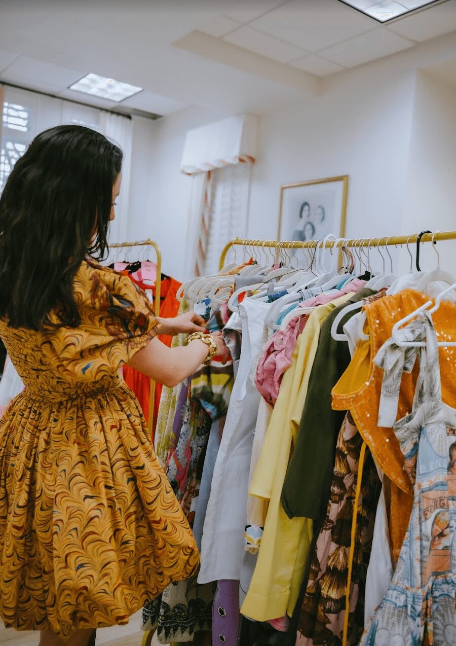 Woman browsing colorful clothes on a rack in a store, with white walls and ceiling lights visible.