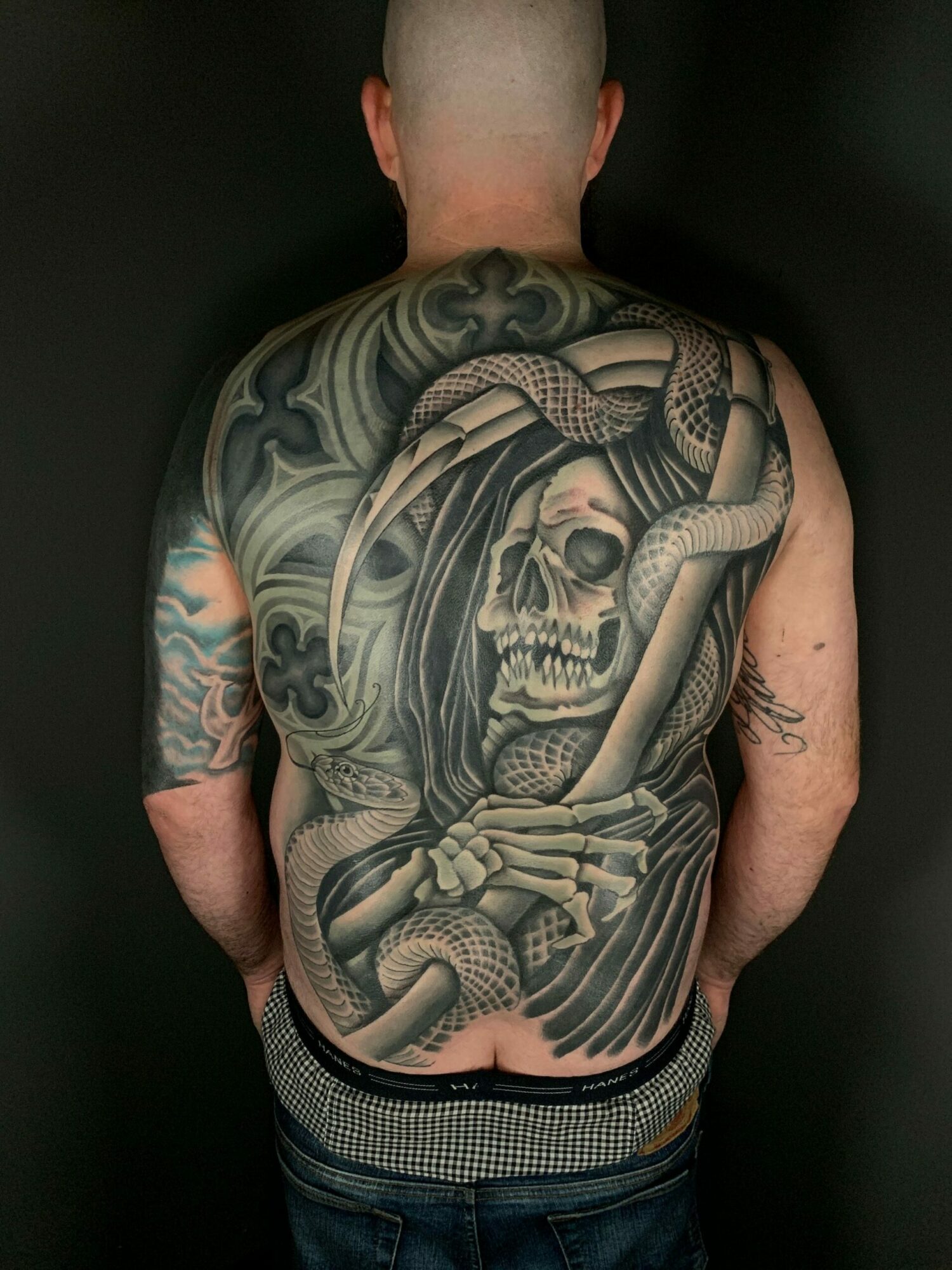 Back view of a person with a large, detailed black and gray skull and snake tattoo covering the shoulder and upper back.