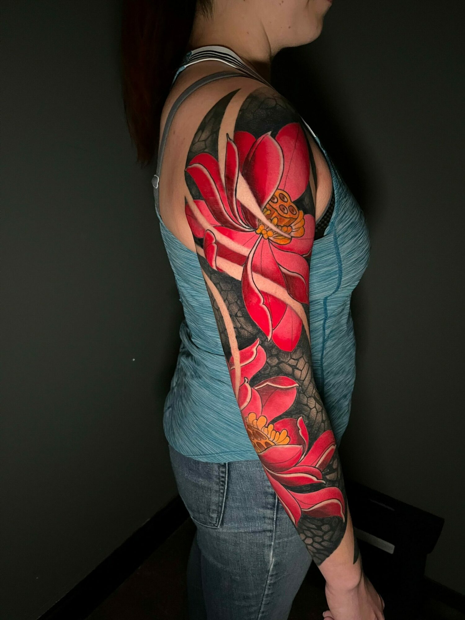 Person lying sideways with colorful tattoo sleeve on arm, wearing blue shirt and jeans, against dark background.
