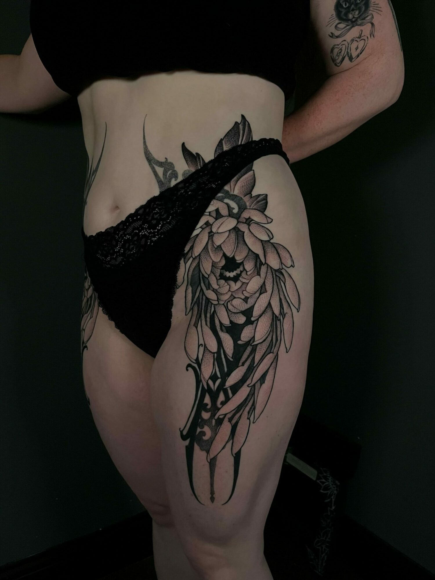 Person with a large floral tattoo on their thigh, wearing black lace underwear, in a dark setting.