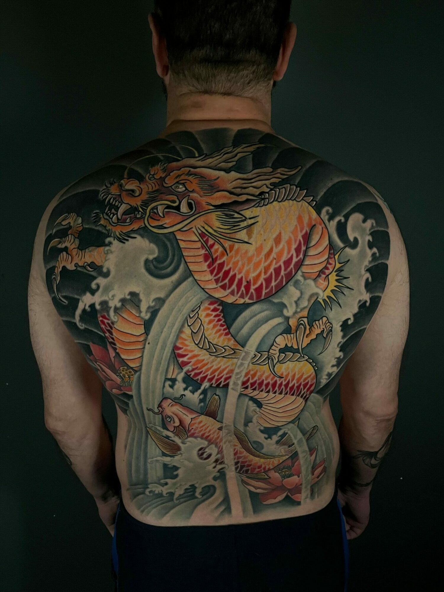 Back tattoo of a dragon with vibrant colors, detailed scales, and surrounding waves, covering the person's upper back and shoulders.