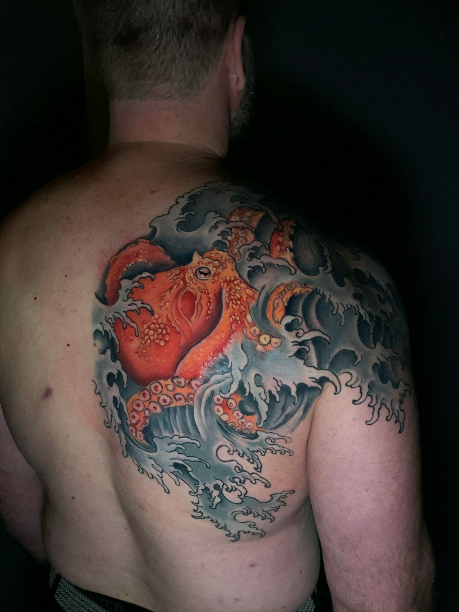 Person with a large, colorful octopus tattoo on shoulder and upper arm, surrounded by waves, on a dark background.