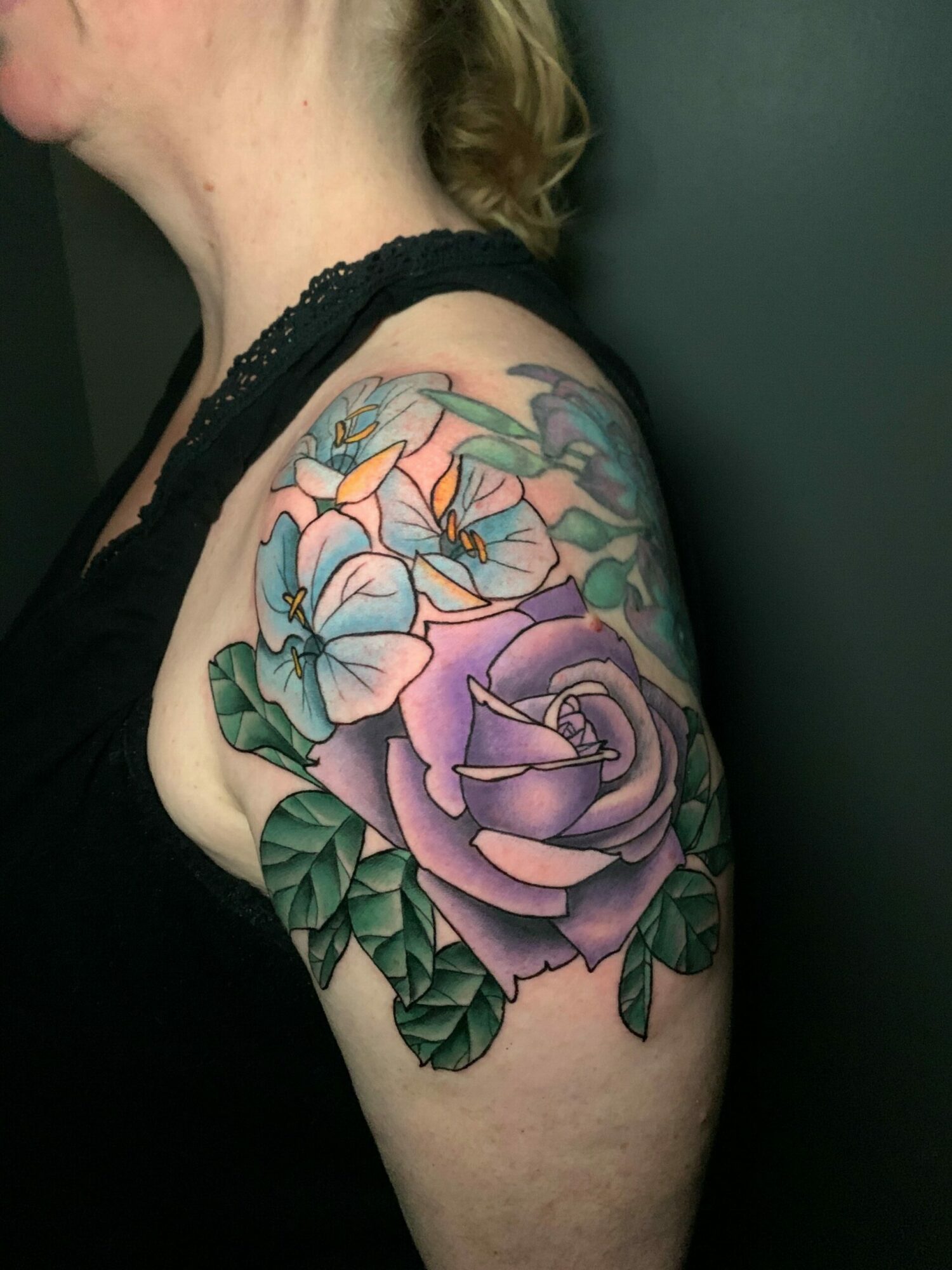 Person with a large colorful tattoo of purple roses and green leaves on upper arm, wearing a black sleeveless top.
