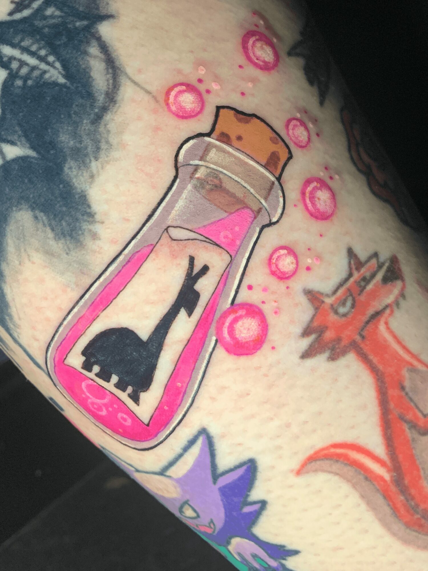 Tattoo of a bottle with a cat silhouette and pink liquid, surrounded by bubbles and other colorful designs.