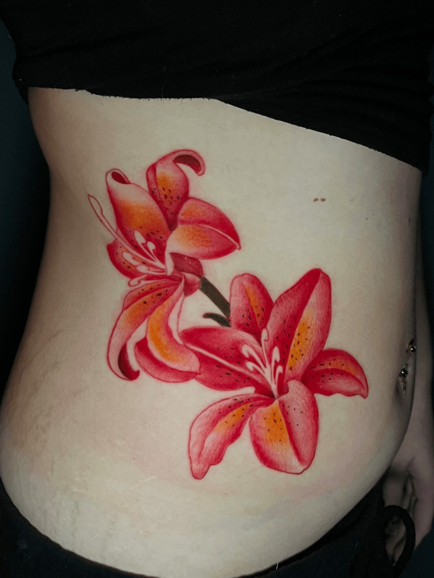 Colorful lily flower tattoo on person's side, with pink and orange petals, and green stem, partially covered by black clothing.