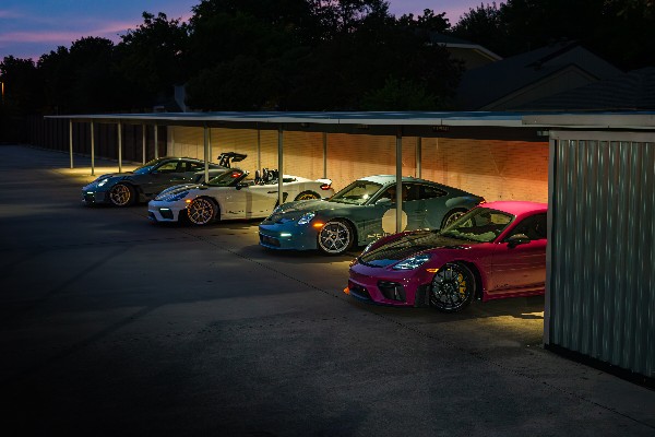 Six sports cars parked under a carport at dusk, illuminated by warm lighting, with trees and houses in the background.