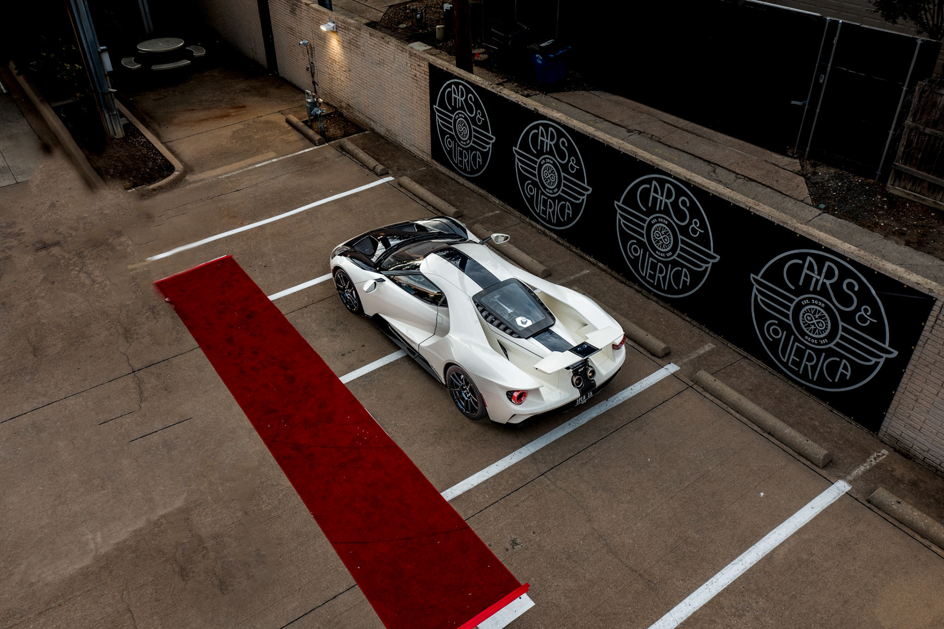 White sports car parked in a parking lot with a red carpet strip nearby, viewed from above.