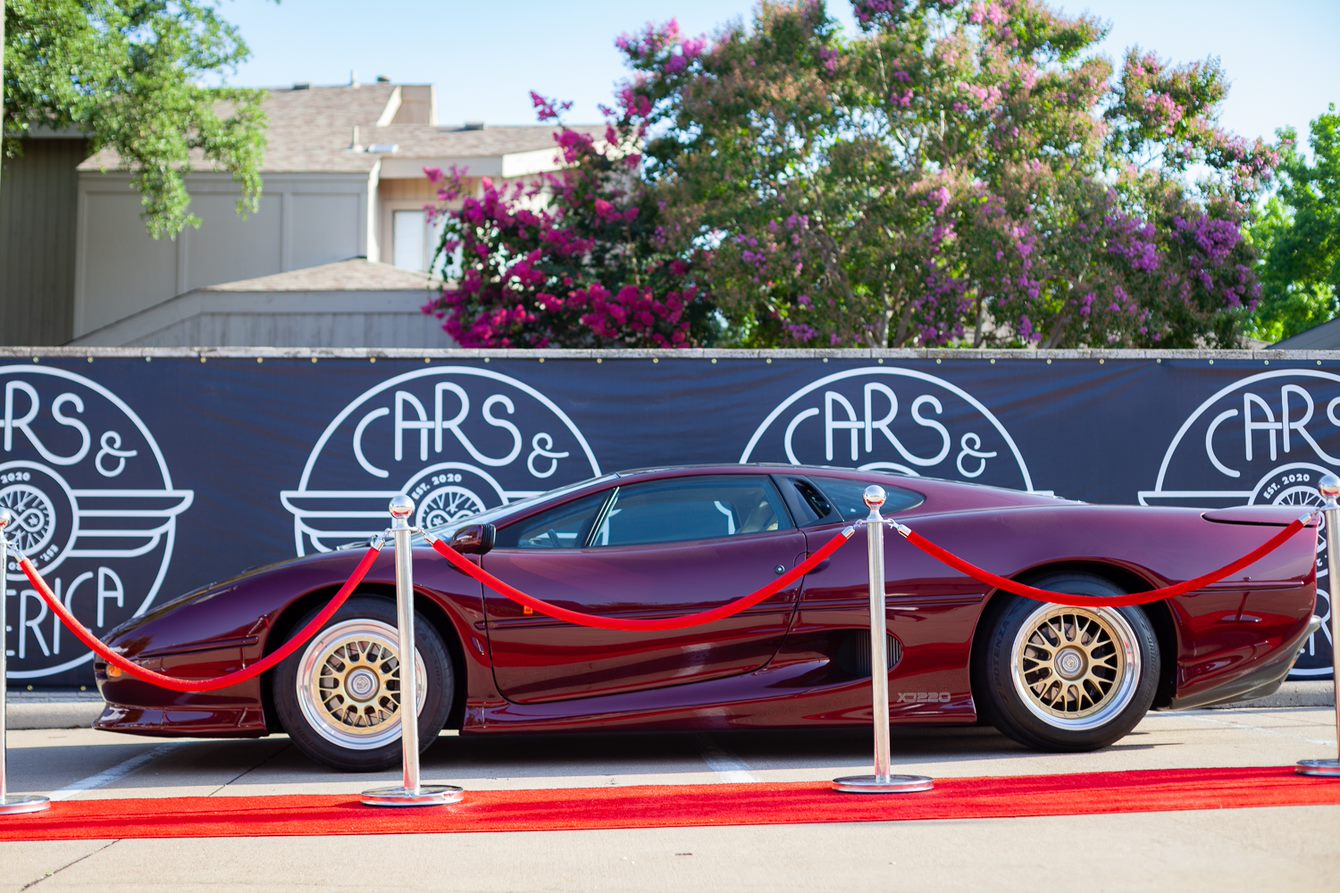 Red sports car displayed behind red ropes, with a black banner and trees in the background.