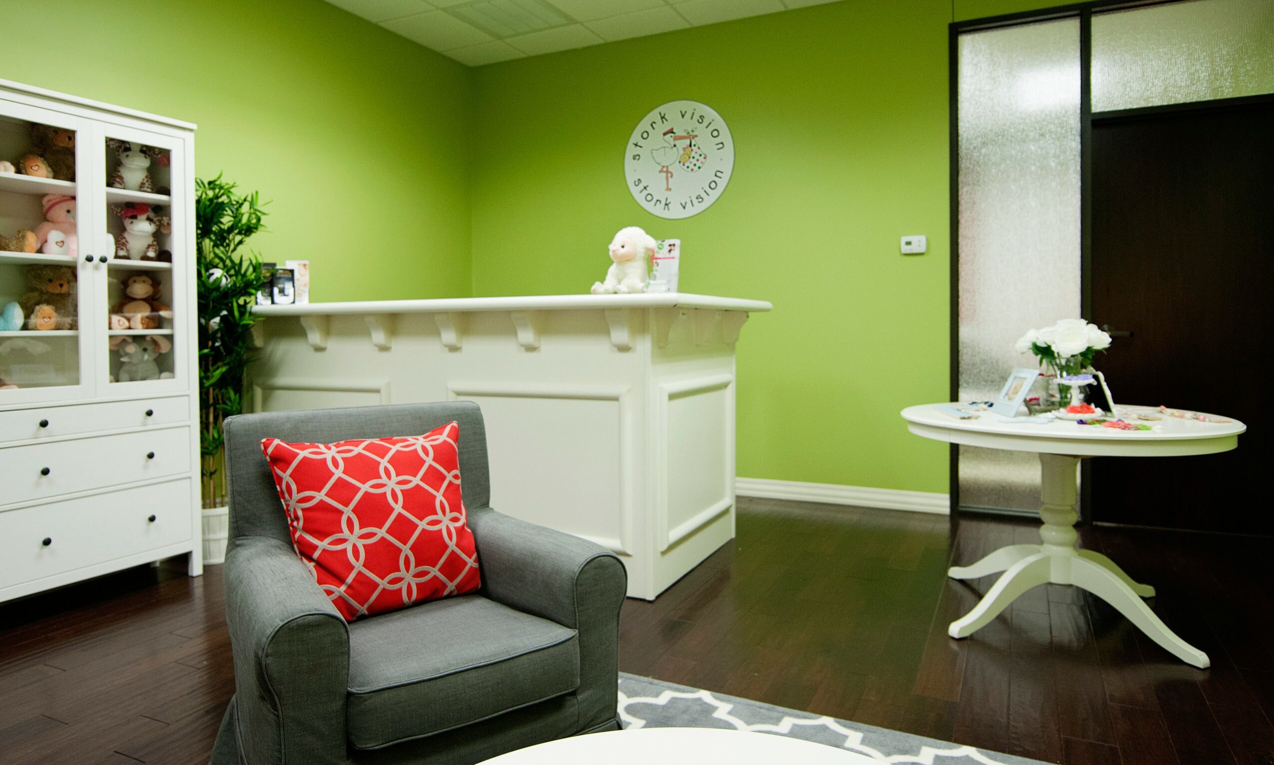 Green-walled room with a gray armchair, red pillow, white cabinet, round table with flowers, and a clock on the wall.