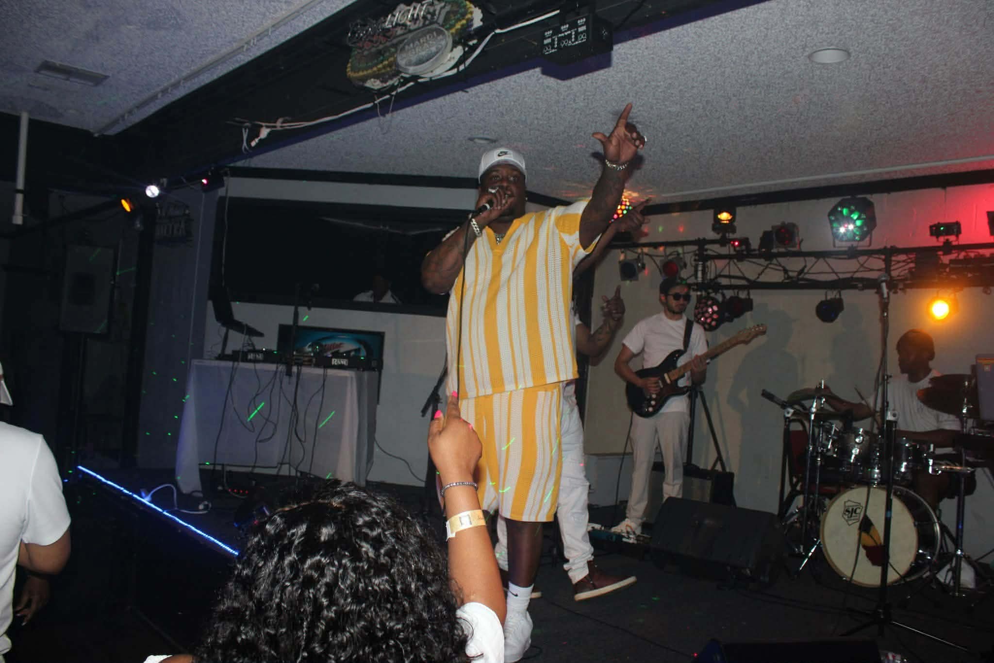 Performer singing on stage with band, raising hand, wearing striped outfit, in a dimly lit venue with colorful lights.