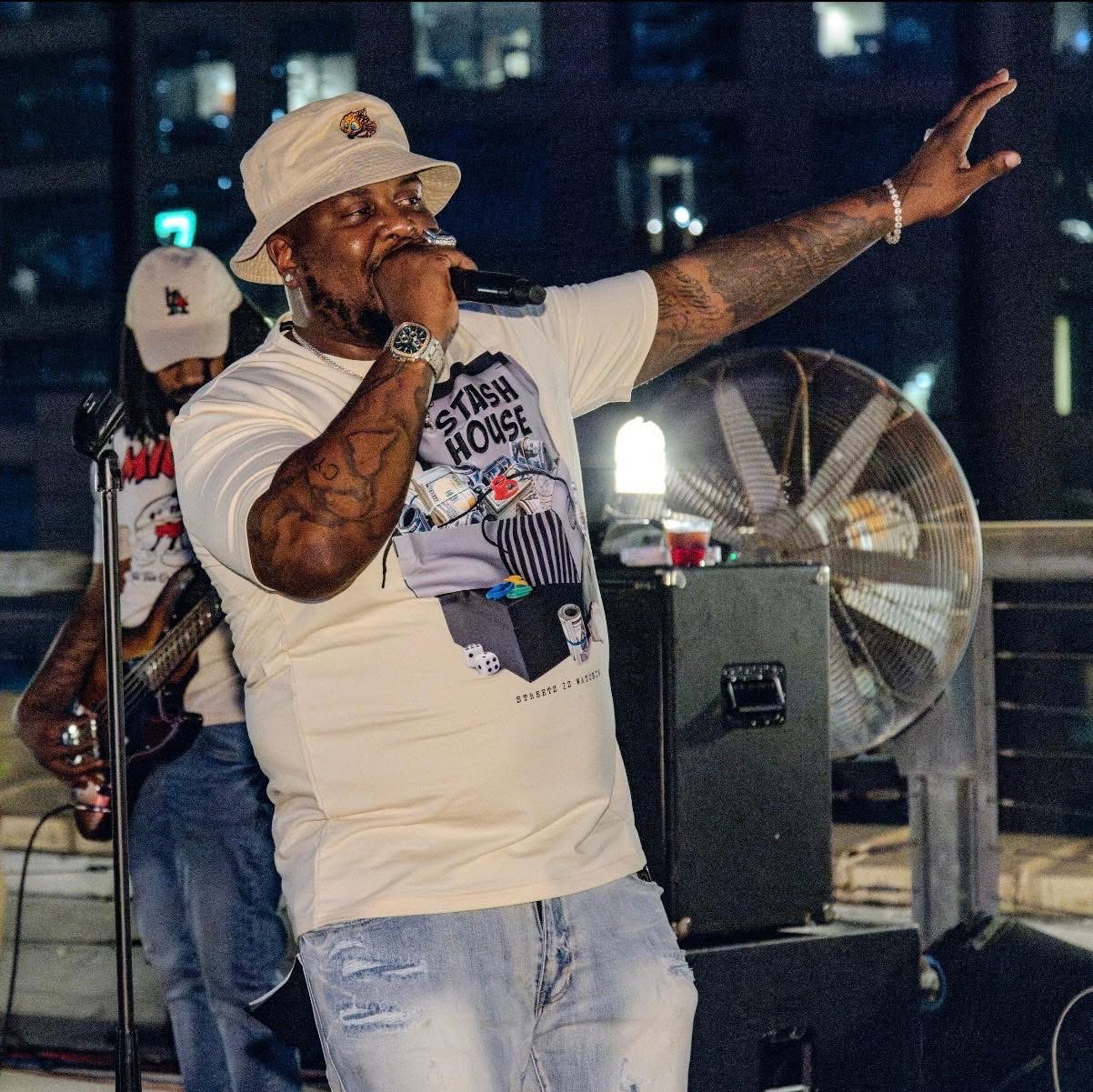 Man singing into microphone with arm raised, wearing a white cap and t-shirt, at night with city lights in background.