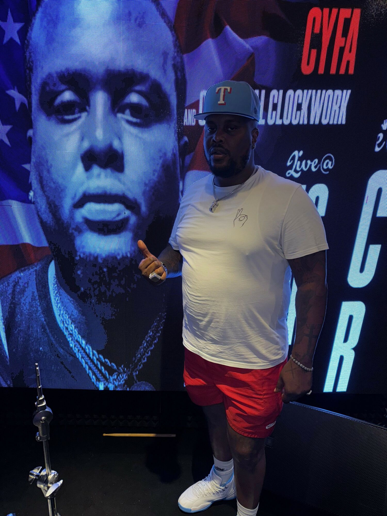 Person in white shirt and red shorts standing in front of a large black-and-white face image on a screen, holding a drink.