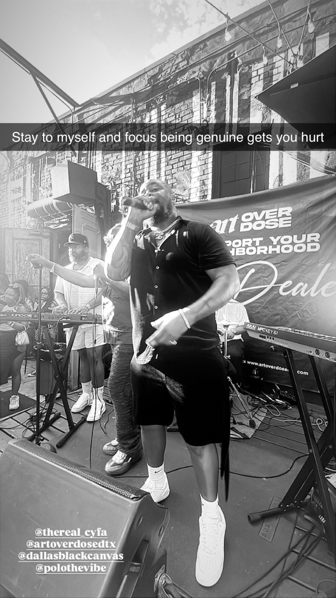 Person standing on stage with musical equipment, wearing a hoodie and shorts, in black and white.