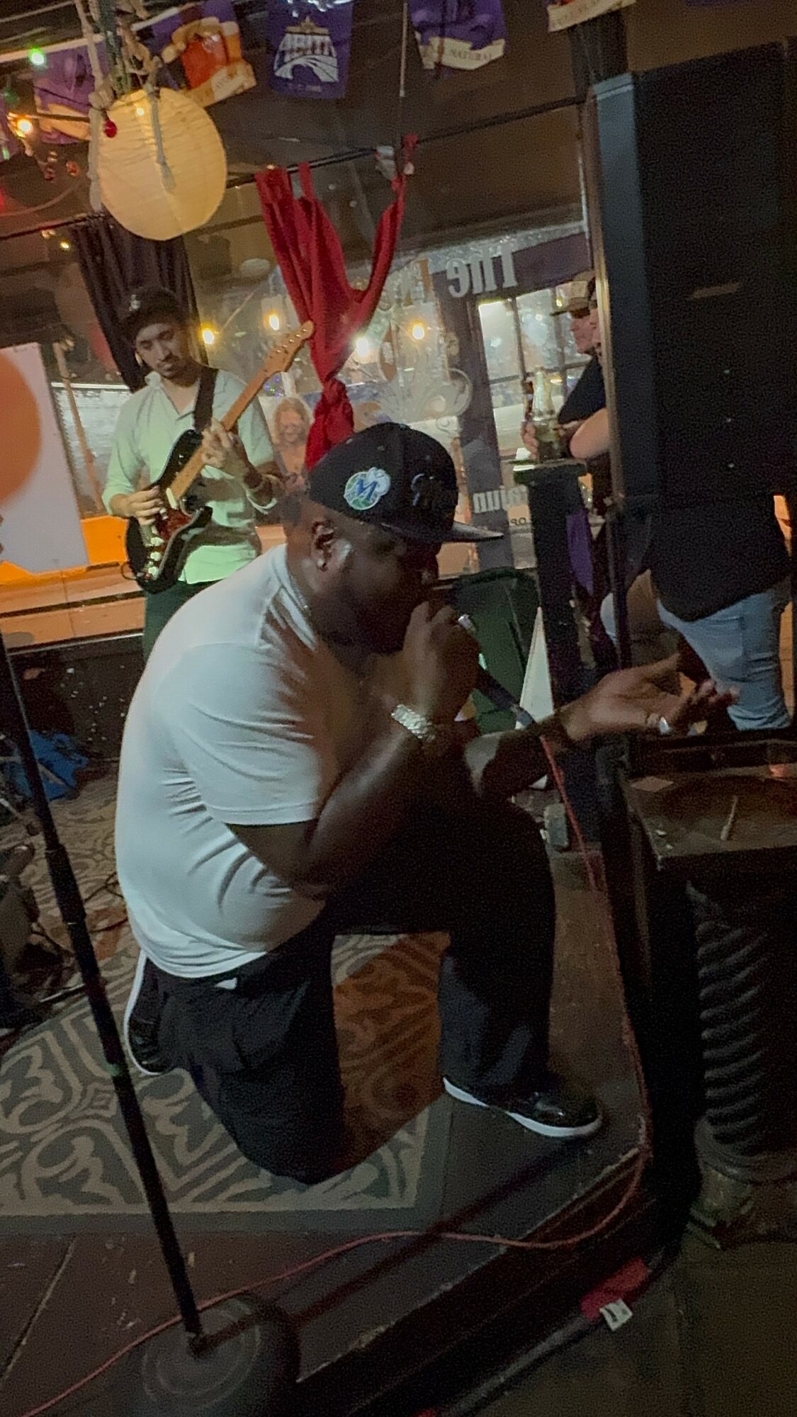 Person sitting on a stage with a microphone, a guitarist in the background, and a guitar hanging from the ceiling.