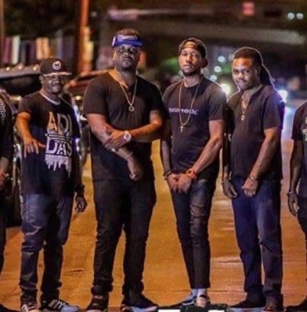 Four men standing outdoors at night, some wearing black clothing and accessories, with blurred city lights in the background.