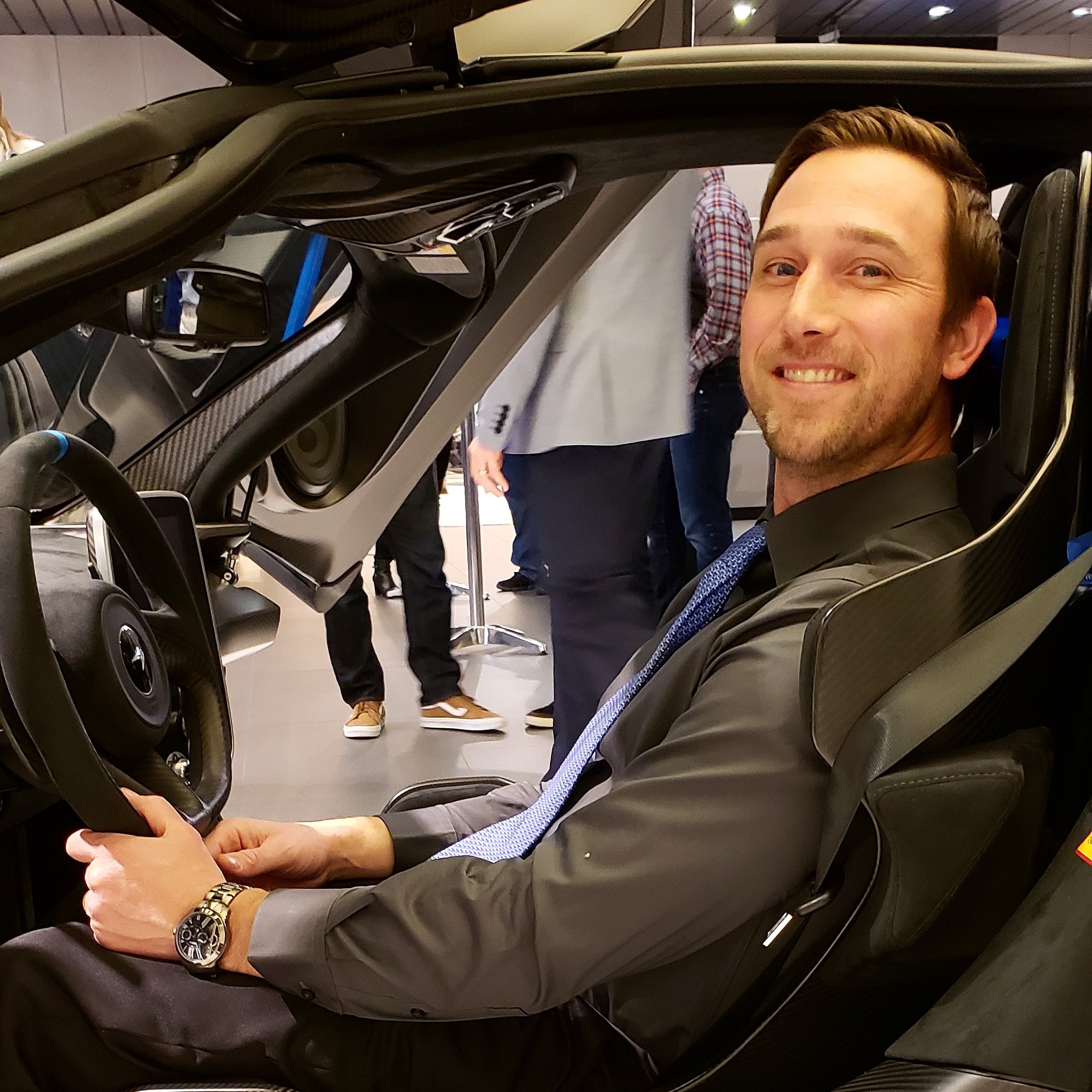 Man sitting in a car, smiling, wearing a black jacket, with people in the background.