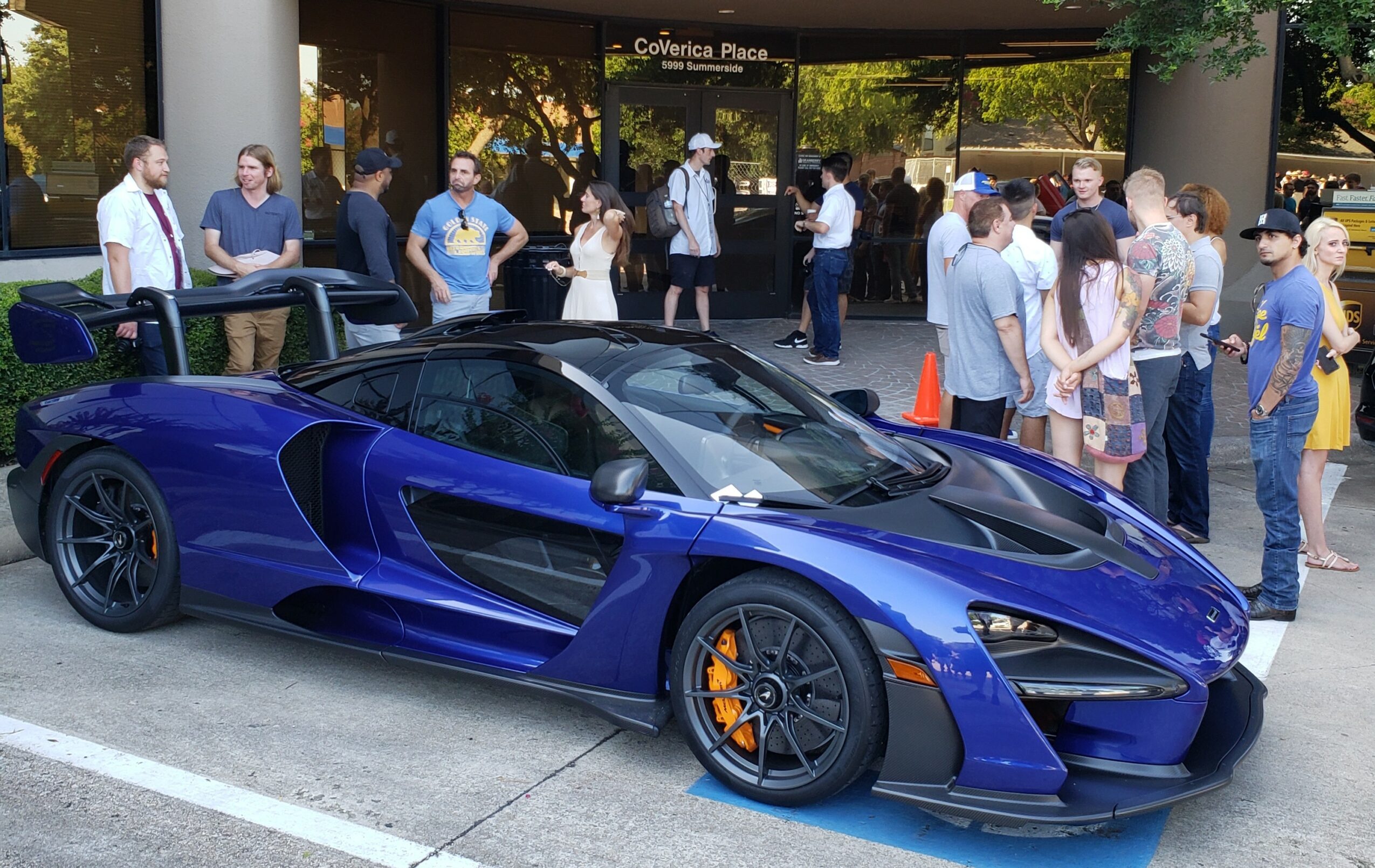 Blue sports car parked on street with people gathered around in background.
