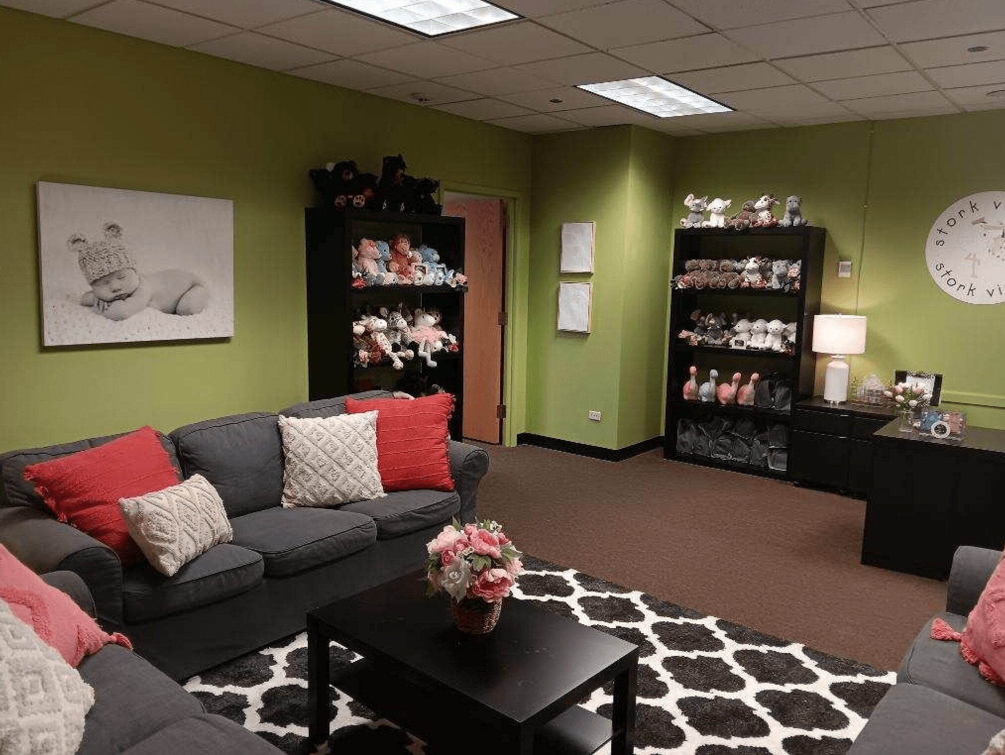 Room with green walls, black shelves filled with plush toys, a gray sofa with red and white pillows, a black coffee table with flowers, and a black desk with a lamp.