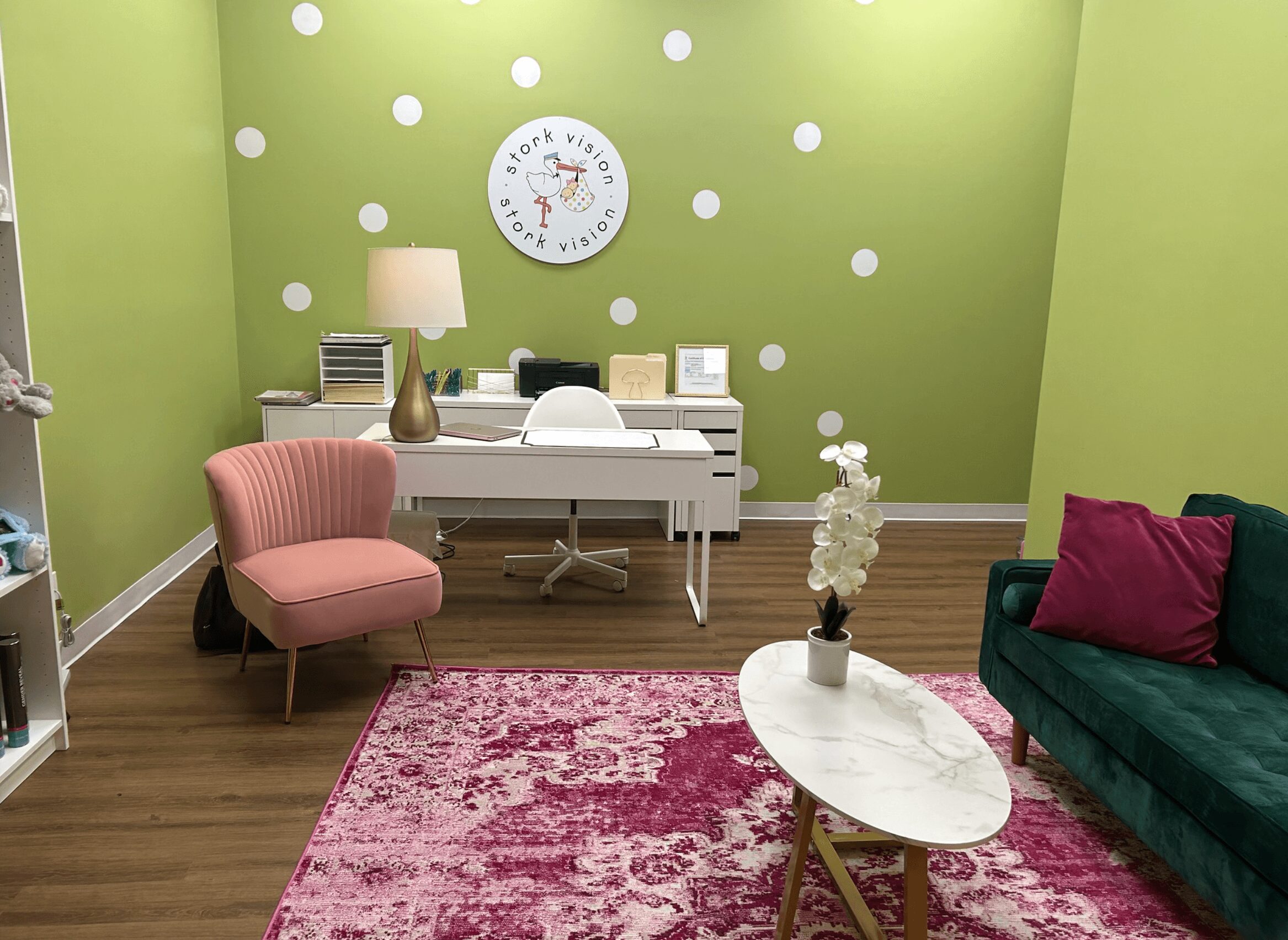 Room with green wall, white desk, pink armchair, green sofa, pink rug, and white clock, decorated with plants and books.