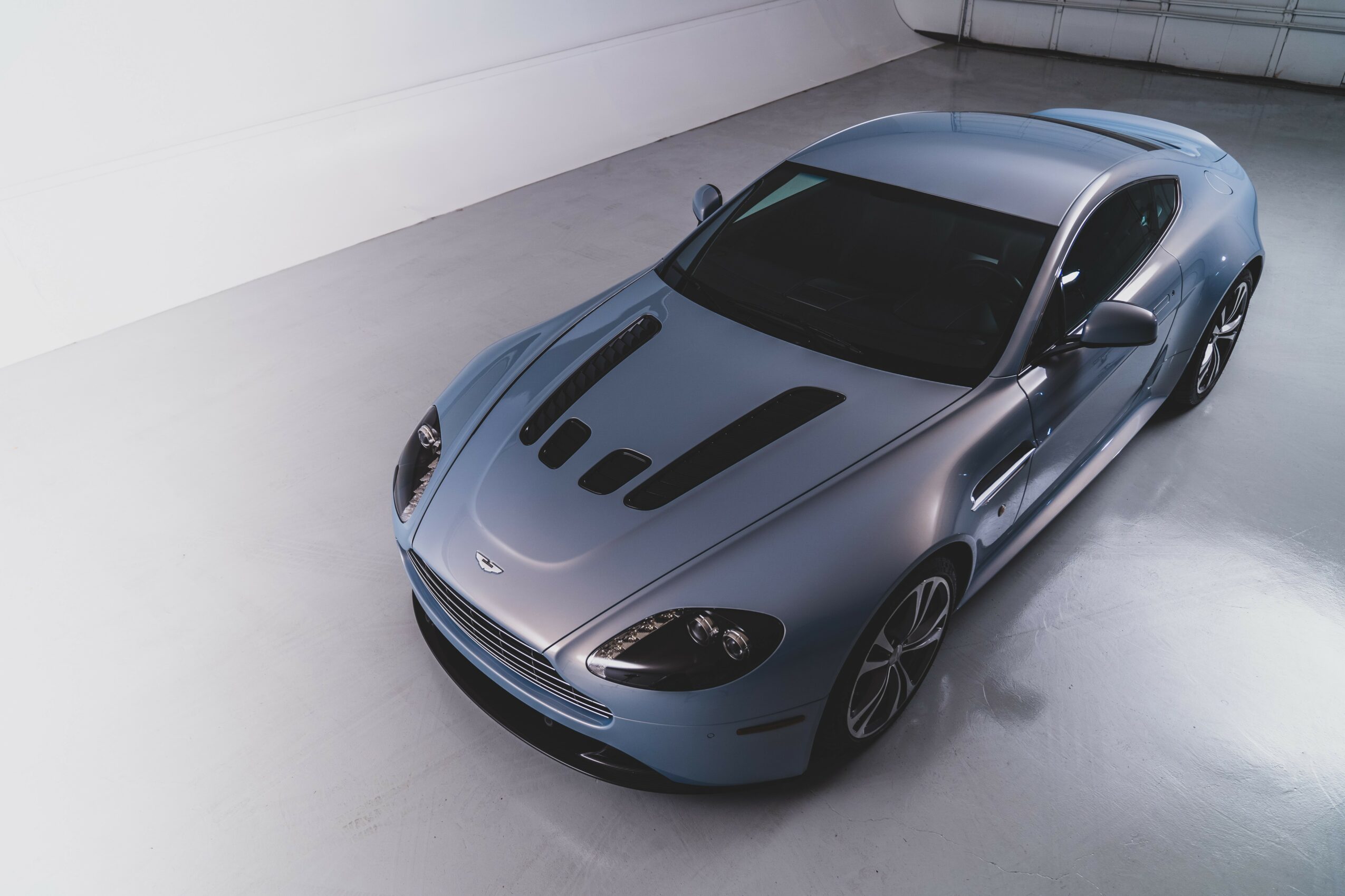 Silver sports car with black racing stripes parked indoors, viewed from above, in a clean, minimal space.